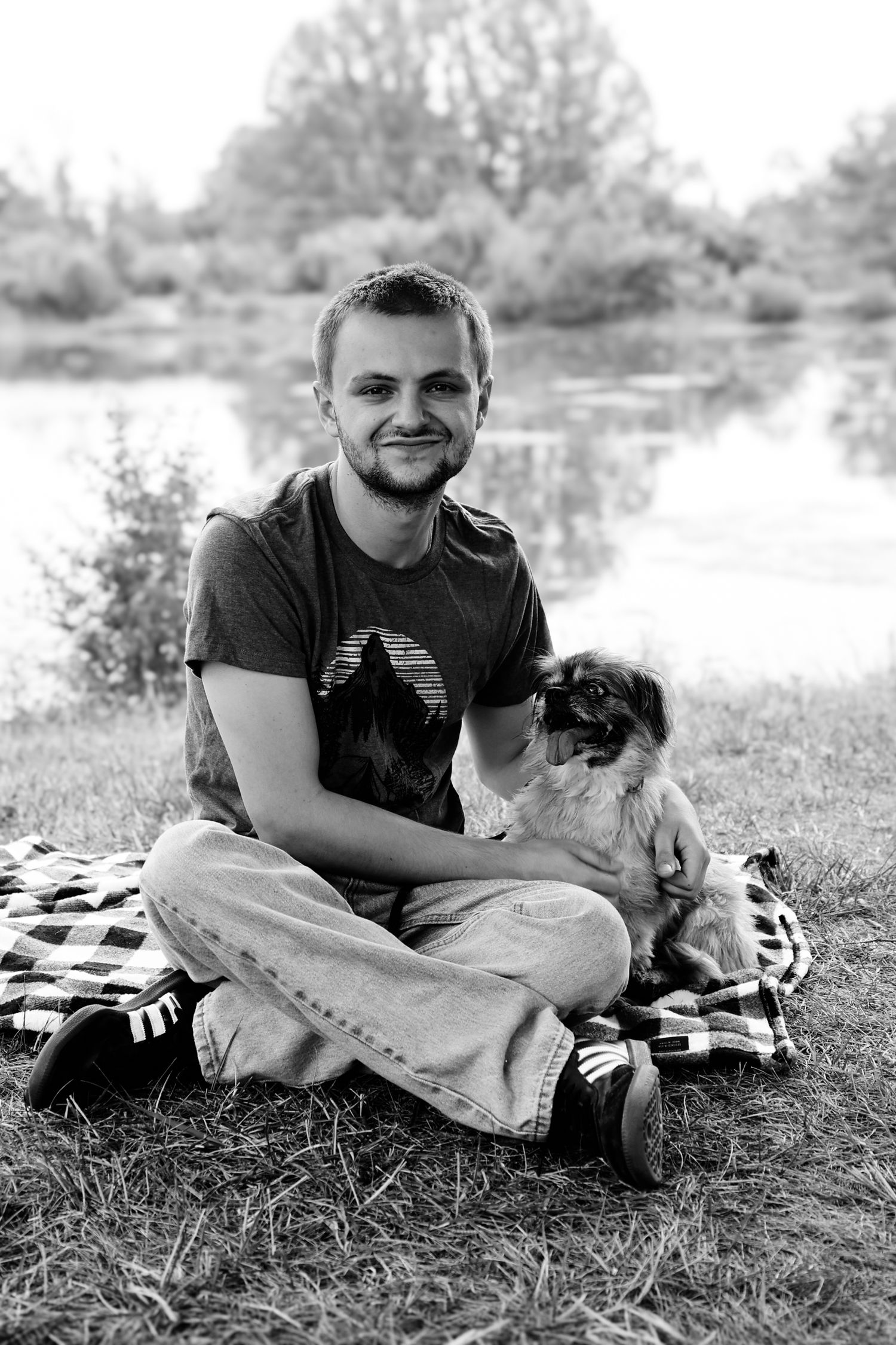 Black and white photo of someone sitting on a blanket by a lake with their small dog on a sunny day.