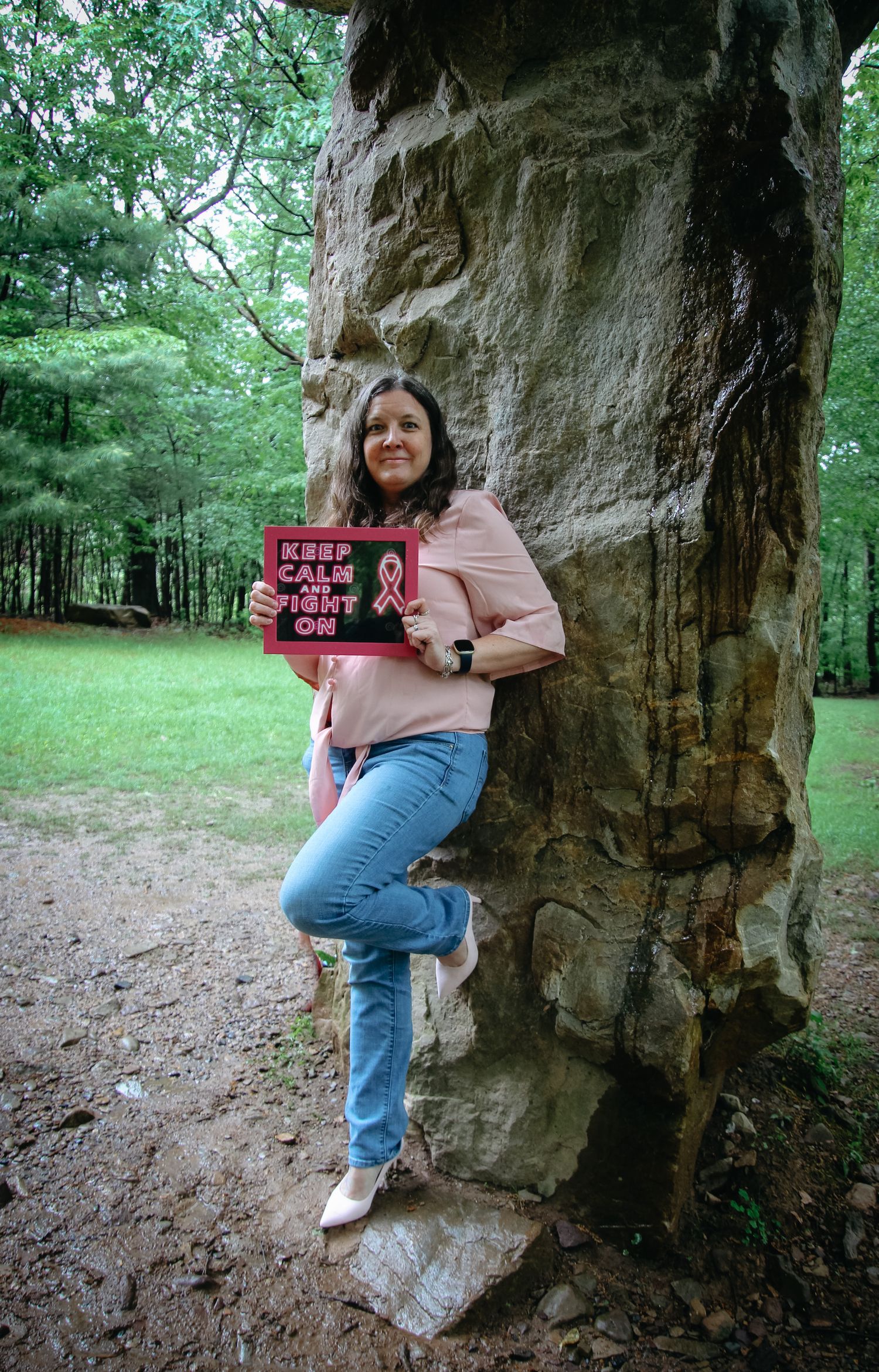 Someone poses against a large rock formation in a green forest setting while holding a sign.