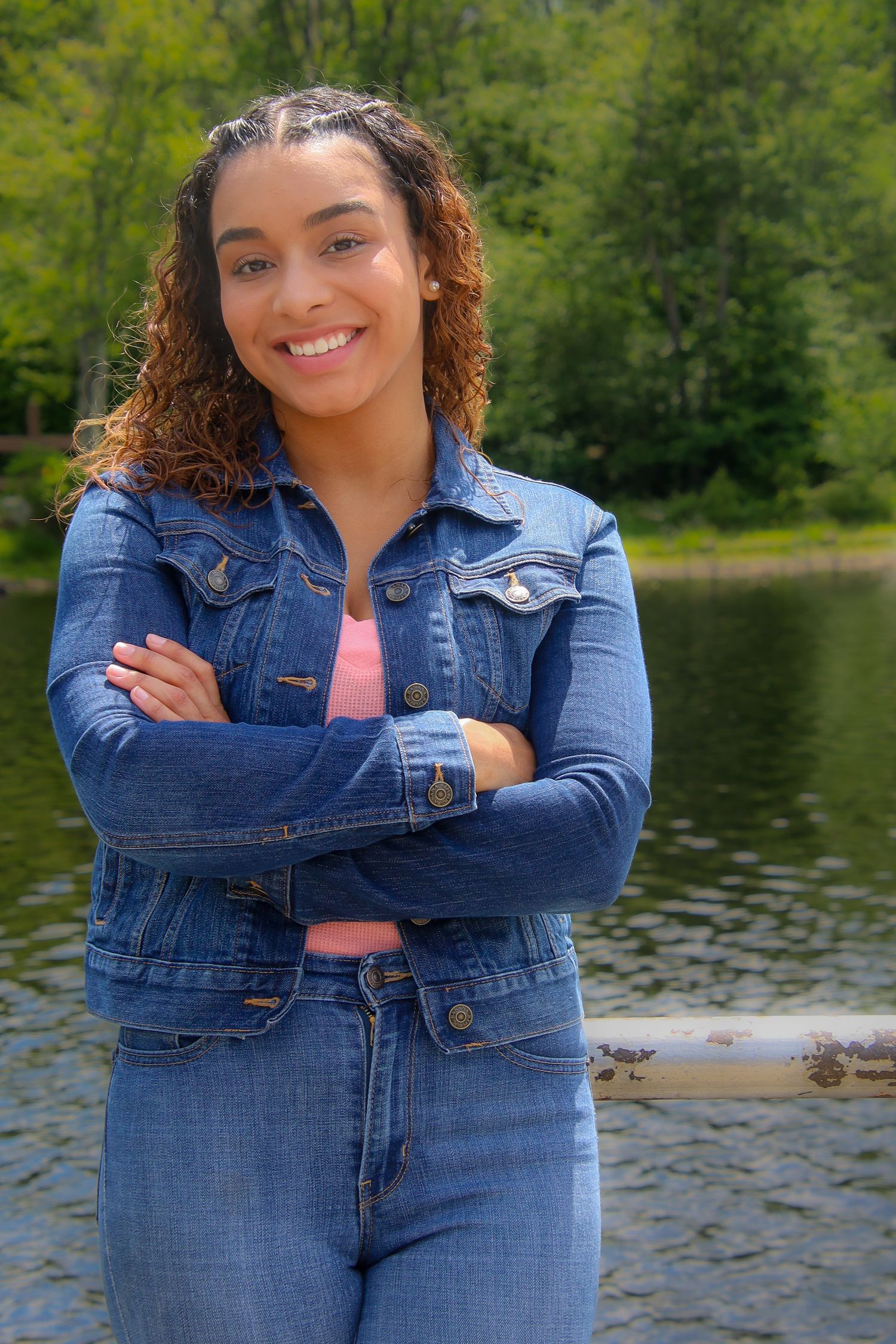 A person in a denim jacket poses confidently by a serene lake with green trees in the background.