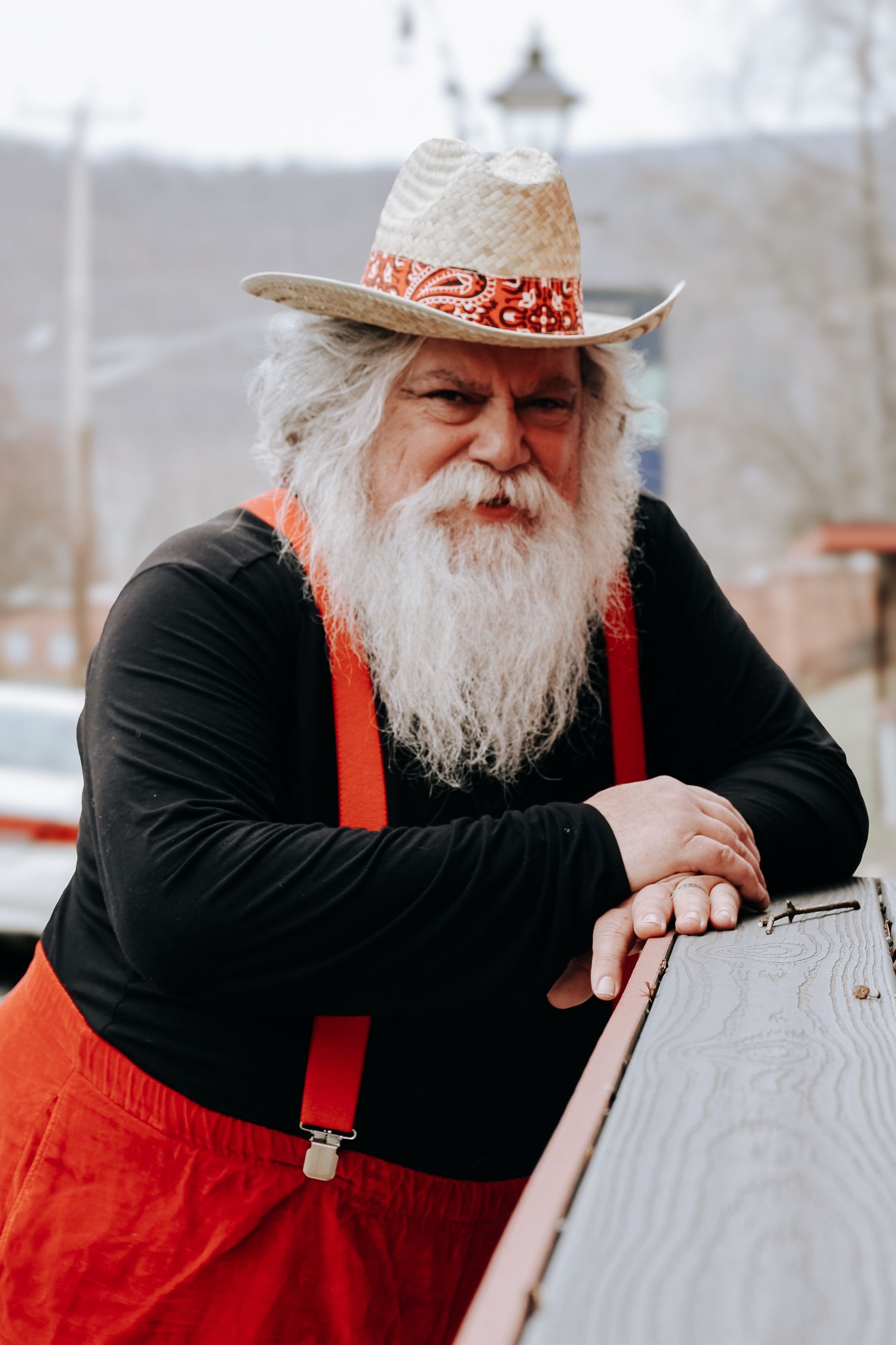A person wearing red overalls and a white hat with a long white beard rests against a railing on a winter day.