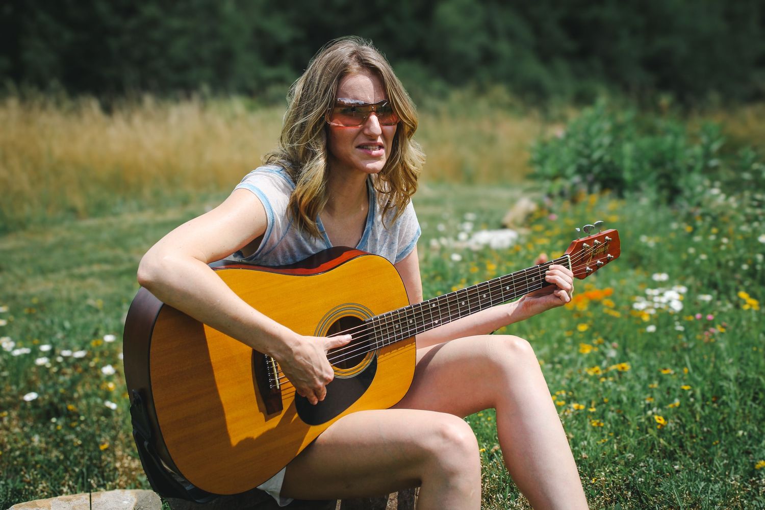 A person in sunglasses plays an acoustic guitar while sitting in a wildflower meadow on a sunny day.