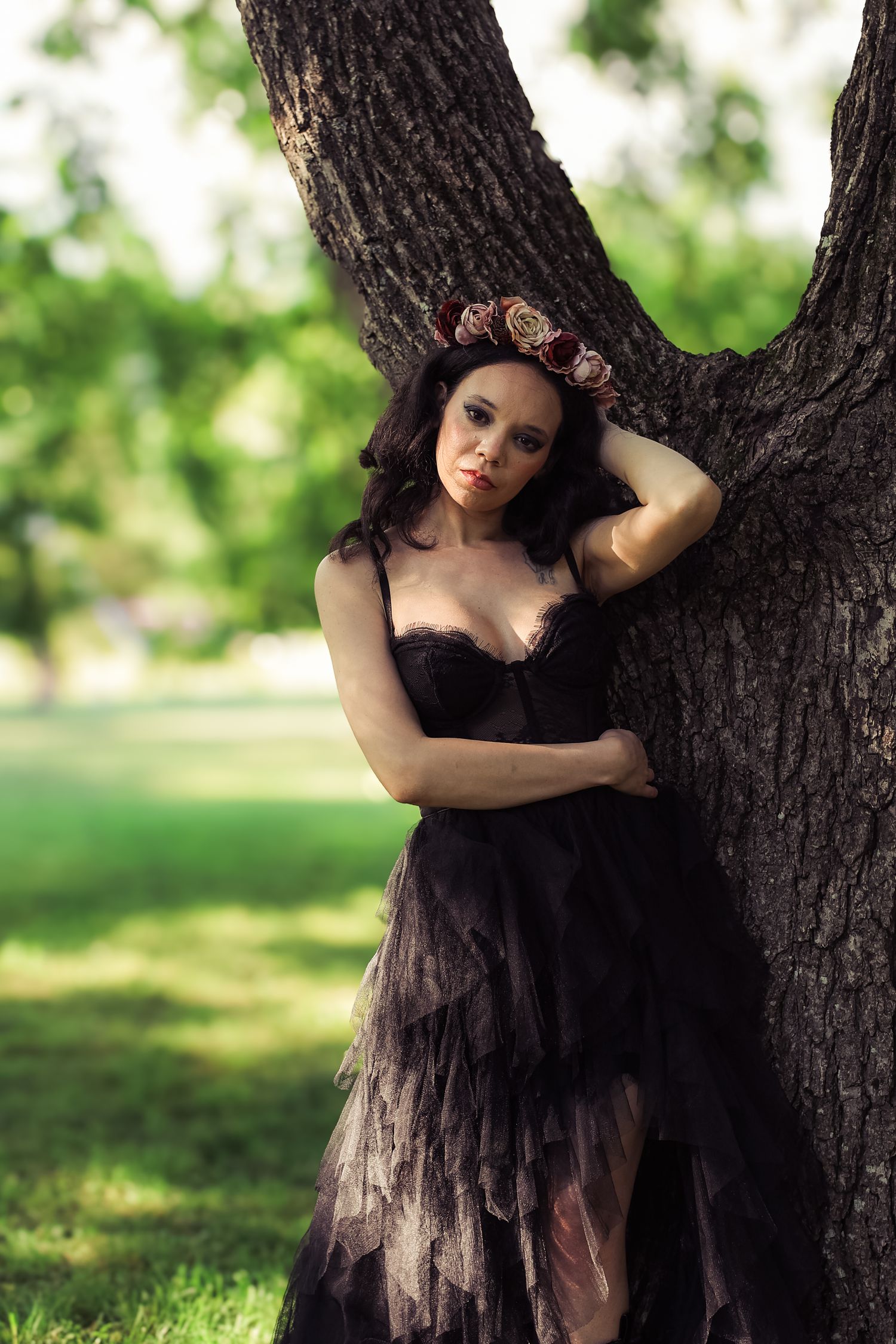 A figure in a flowing black dress poses gracefully against a large tree trunk in a sunlit outdoor setting.