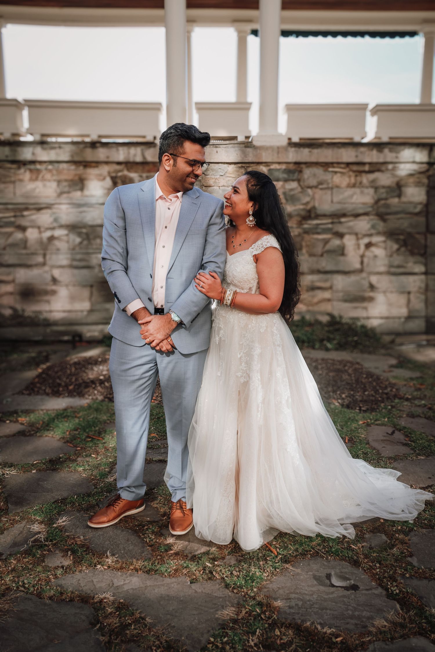 A romantic wedding couple poses together against a rustic stone wall in elegant attire.