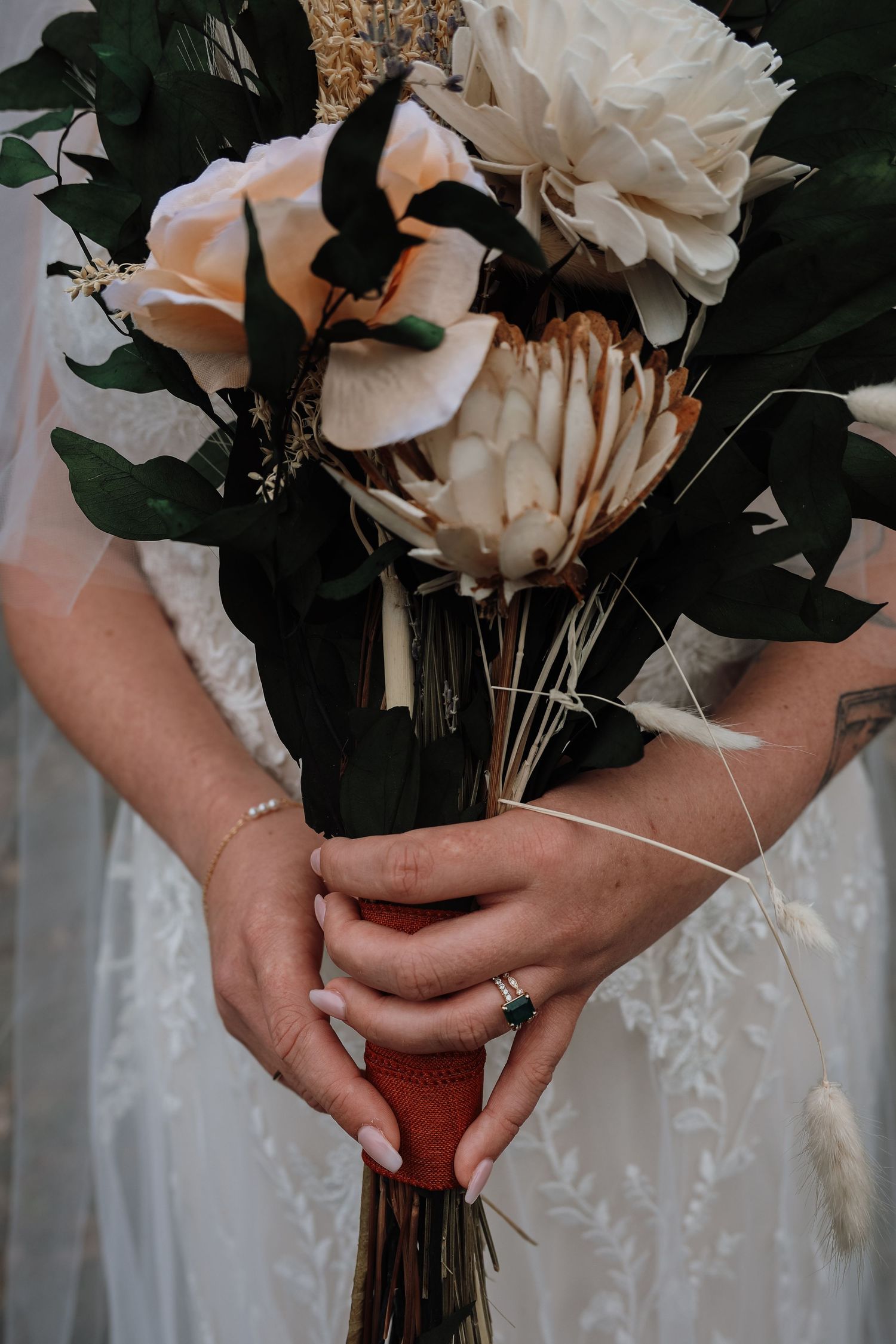 Close-up view of hands holding a bridal bouquet with white protea flowers and dark green foliage against a white dress.