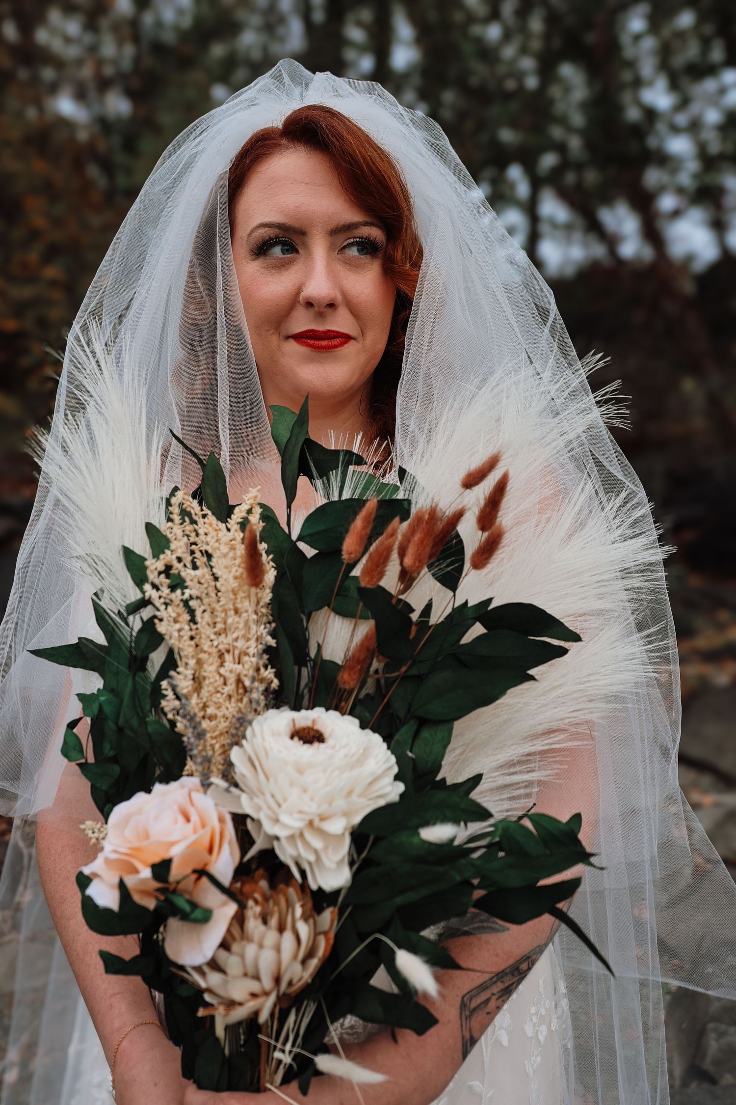 Wedding bouquet featuring white protea, greenery and dried flowers held against dark background with flowing veil.