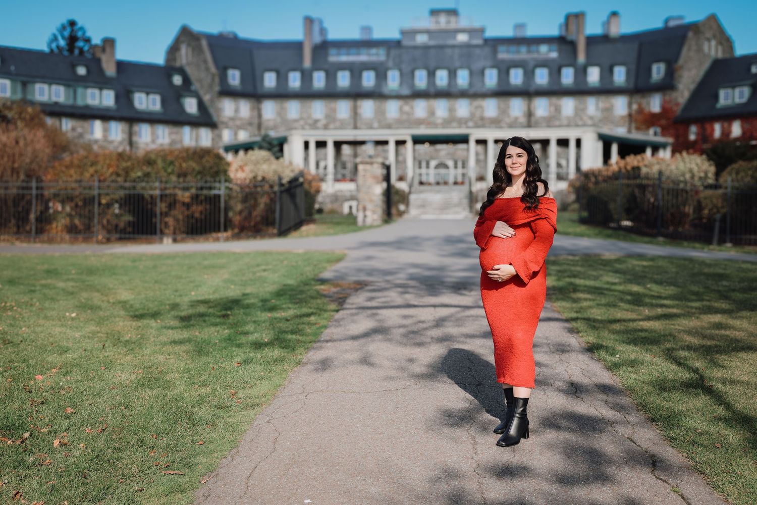Someone in a bright red coat walks along a path leading to a grand historic mansion building on a sunny autumn day.