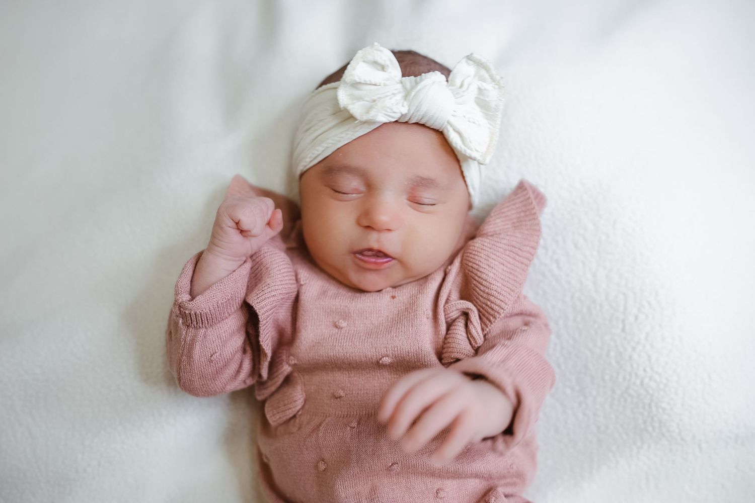 Sleeping newborn baby wearing pink clothing and white hat rests peacefully on soft white blanket.