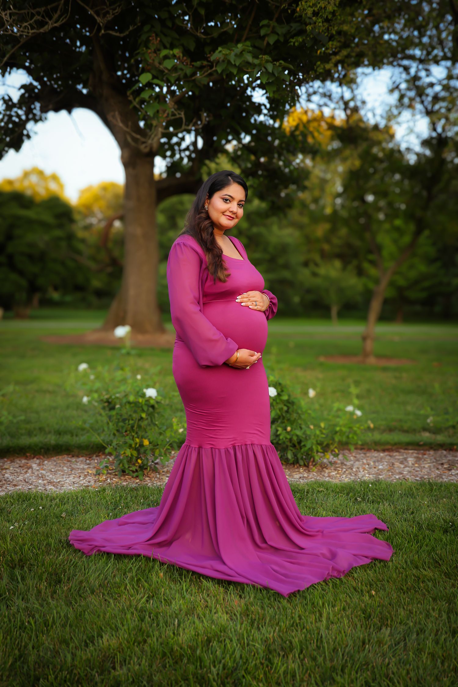 A person in a flowing magenta dress poses gracefully on grass in a park setting during a photo session.