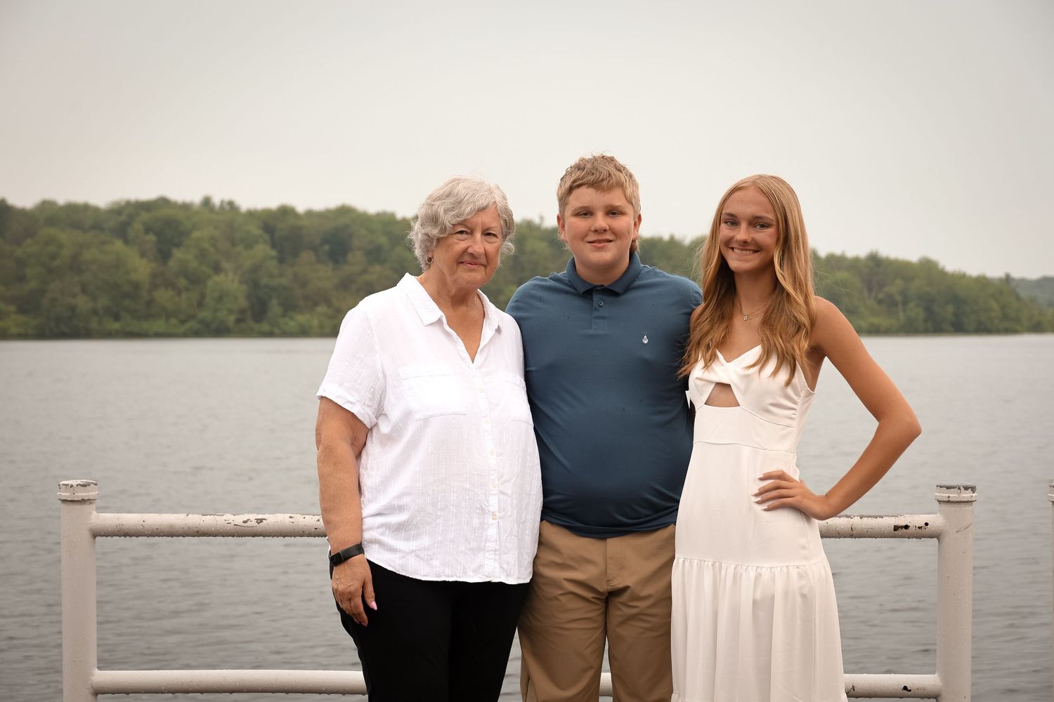 Three people stand together on a wooden dock by a lake on an overcast day.