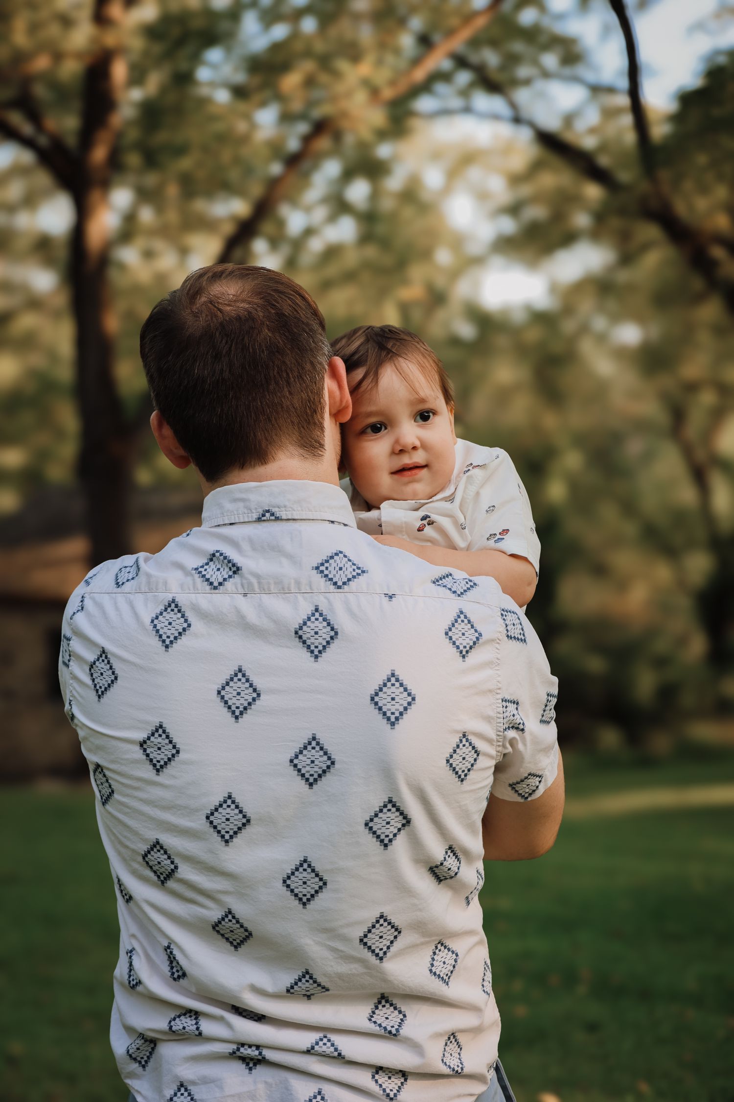 Parent in patterned shirt holds and comforts small child in outdoor park setting during golden hour.