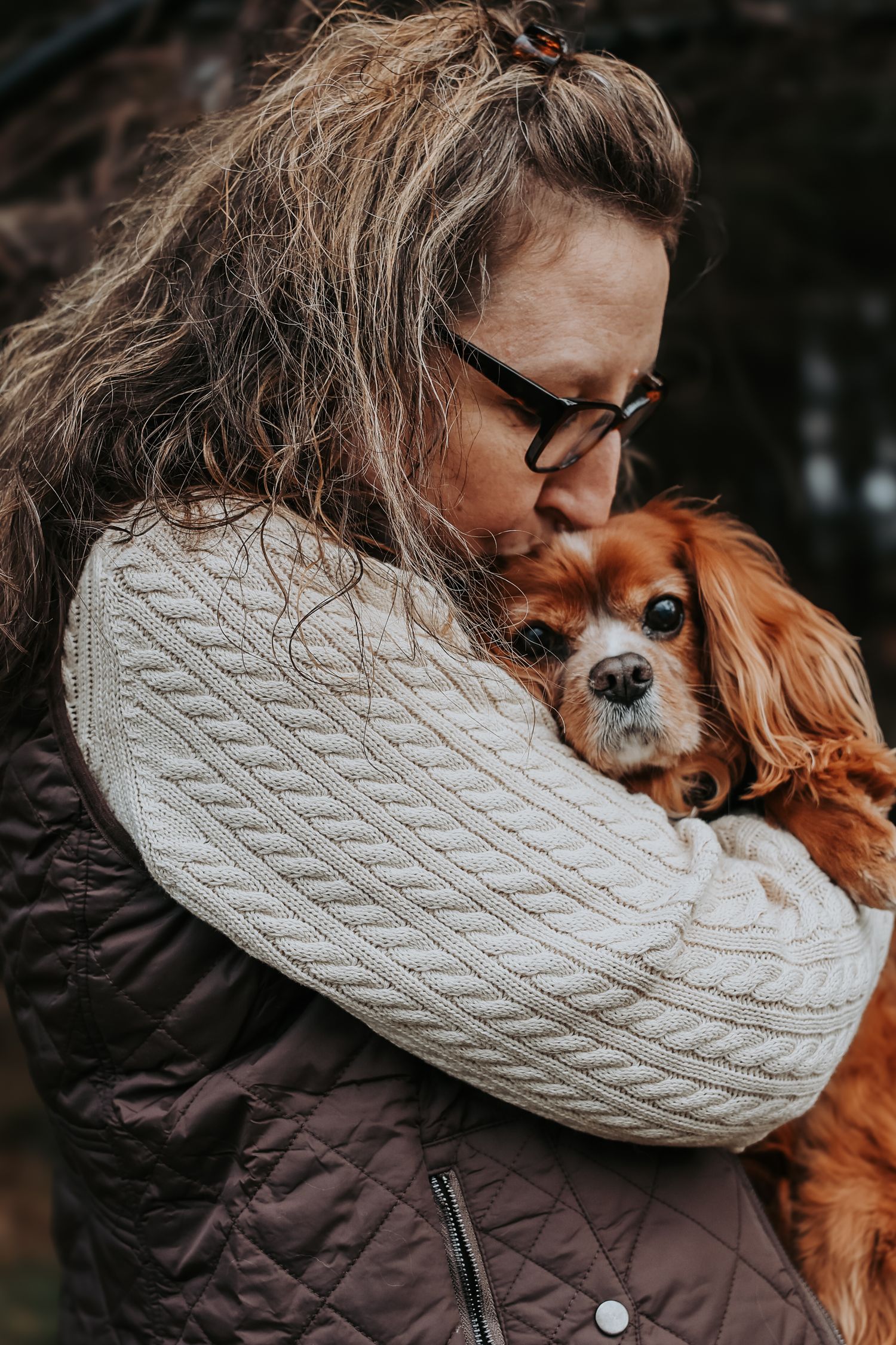 A person in a cable knit sweater and quilted vest hugs their sweet Cavalier King Charles Spaniel dog.