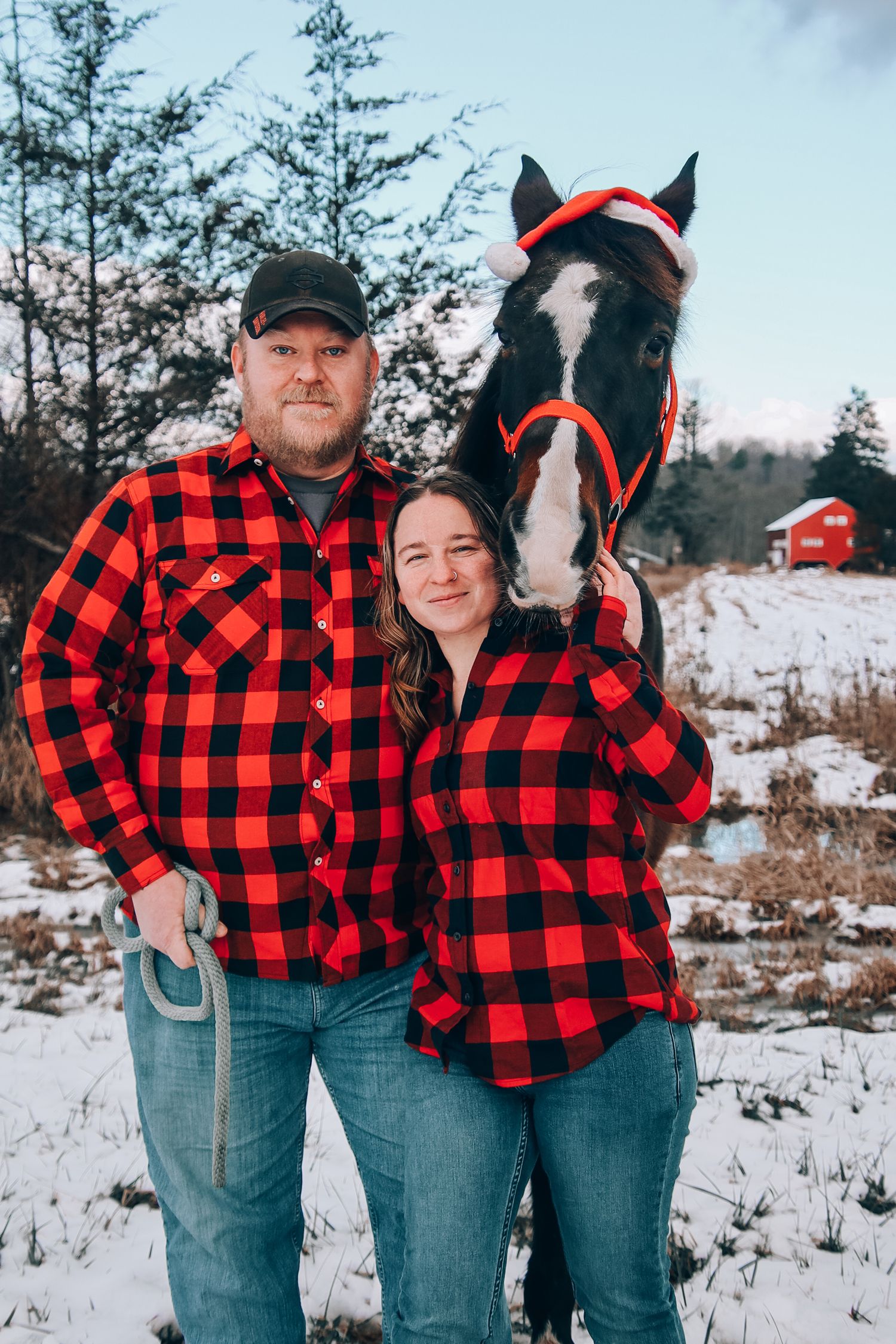 A couple in matching red buffalo plaid flannel shirts poses with their black horse in a snowy winter setting.