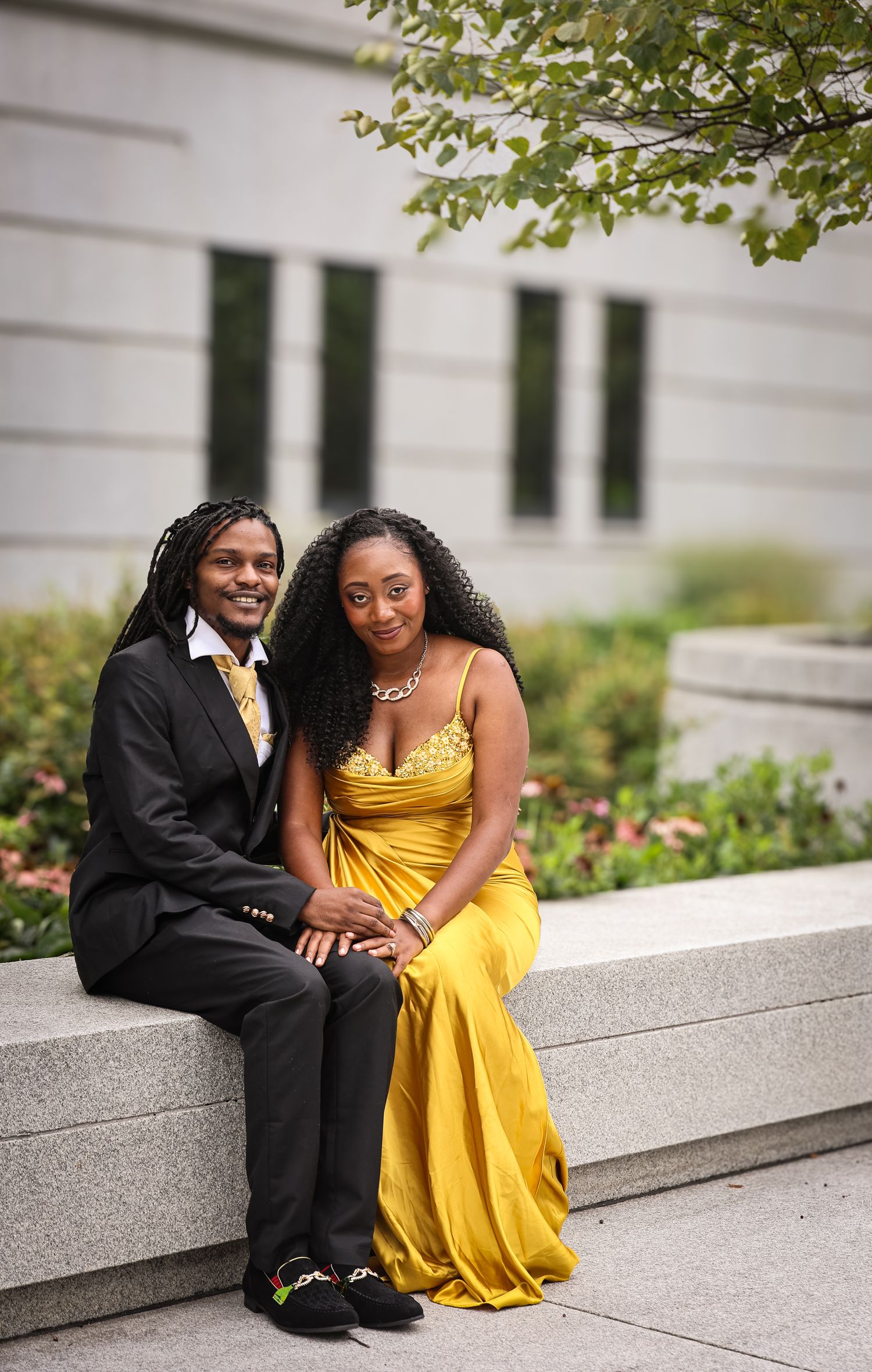 A couple sits together on stone steps, with the person in a bright yellow dress and the other in a black suit.