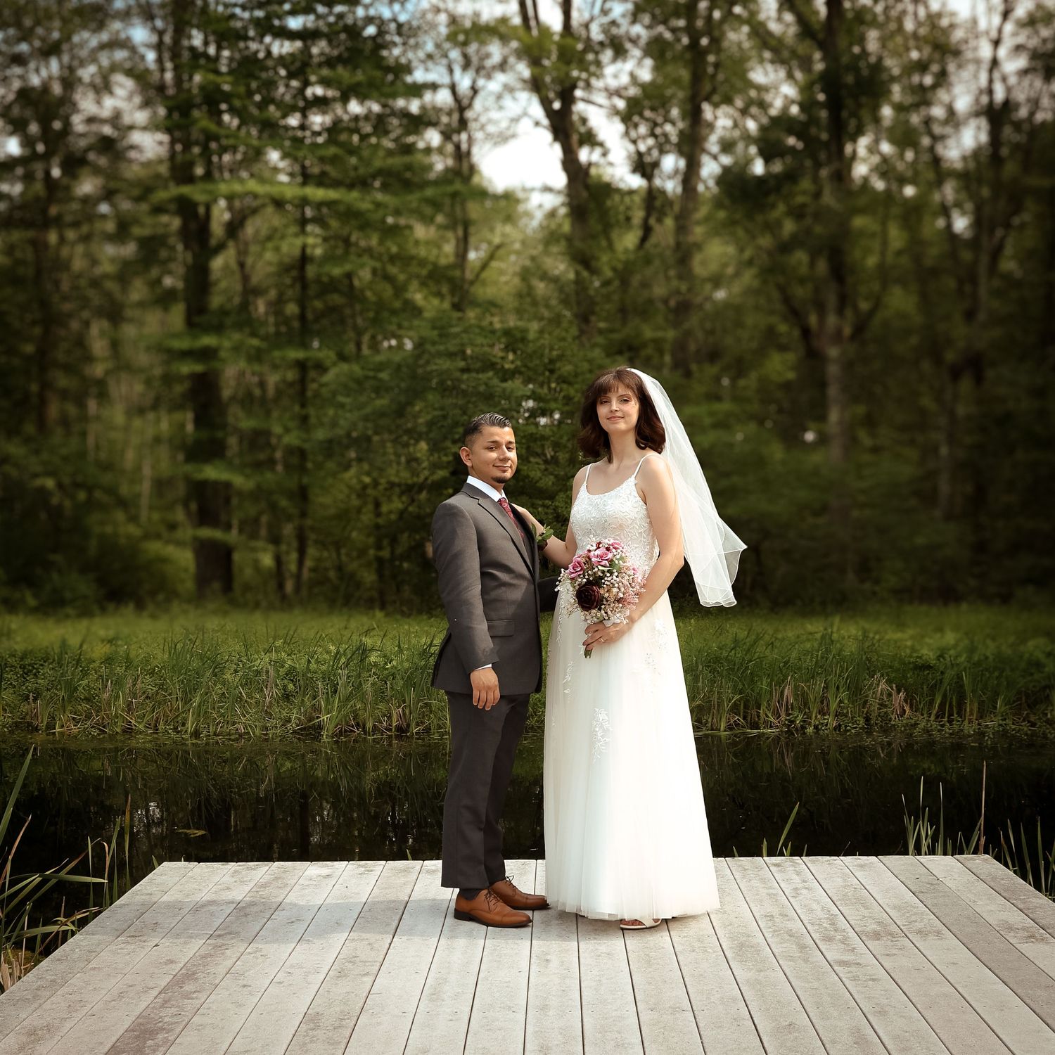 A couple in wedding attire stands on a wooden deck surrounded by lush green trees in a romantic outdoor setting.