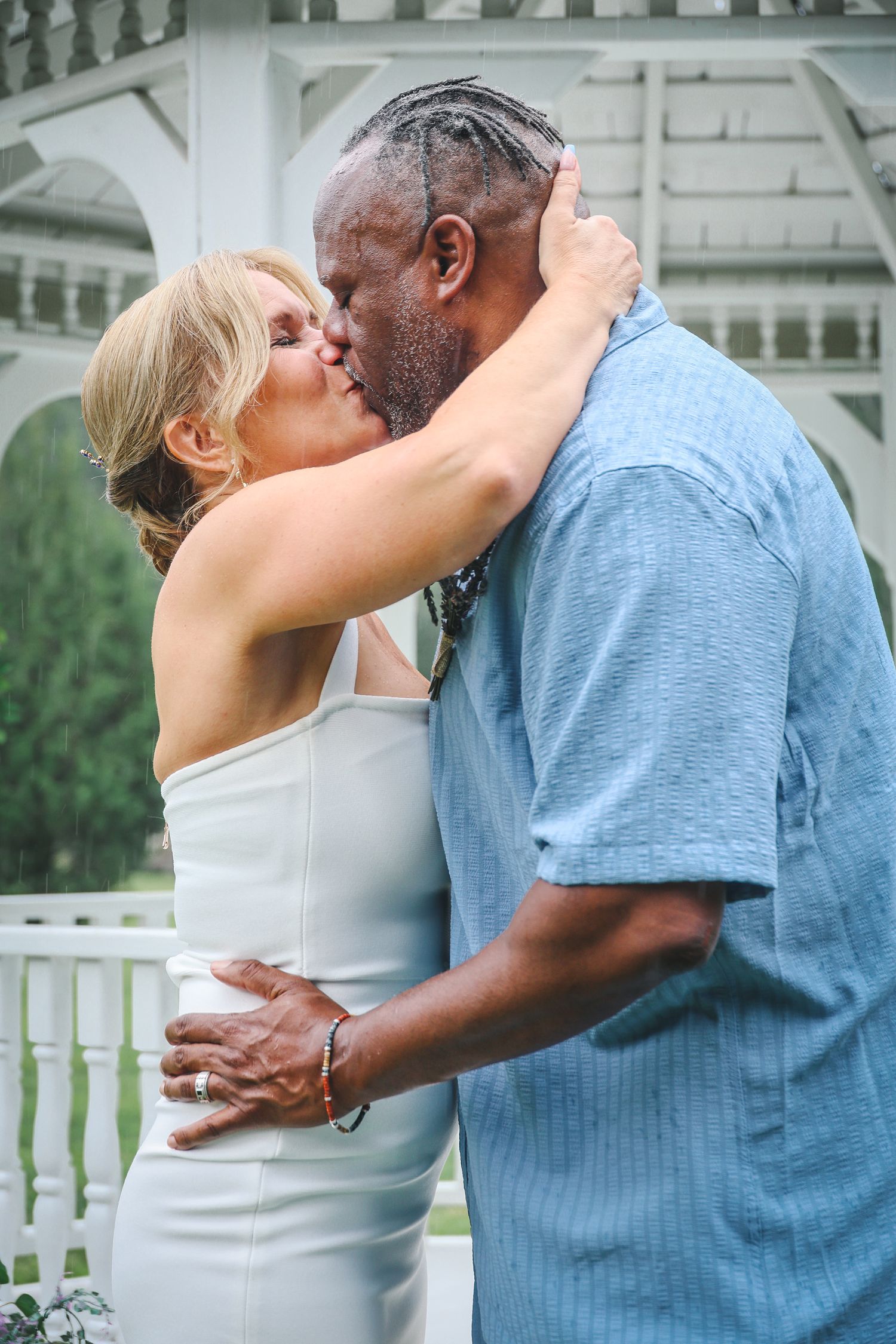 A couple shares a romantic kiss on a porch with a white gazebo in the background.