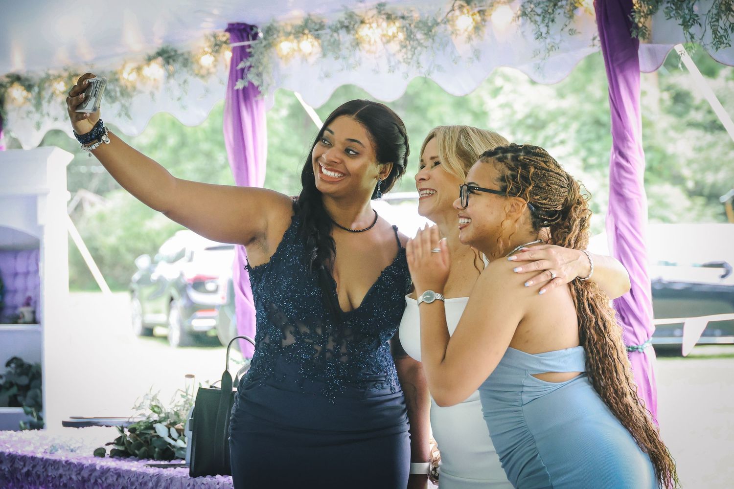 Two people in formal attire take a selfie together under a decorated canopy with purple ribbons at an outdoor event.