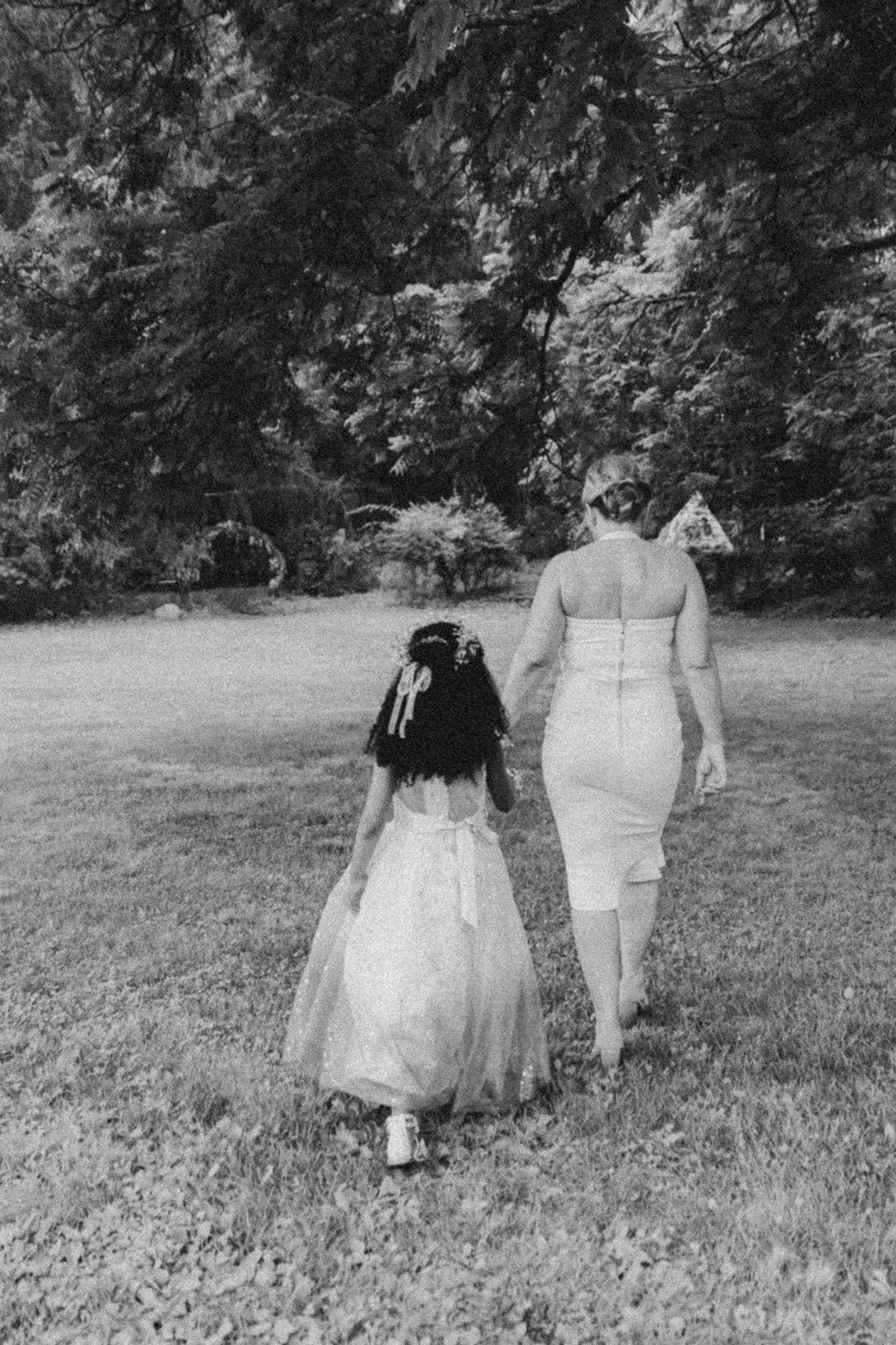 A couple in wedding attire walks away from the camera across a grassy field in black and white.
