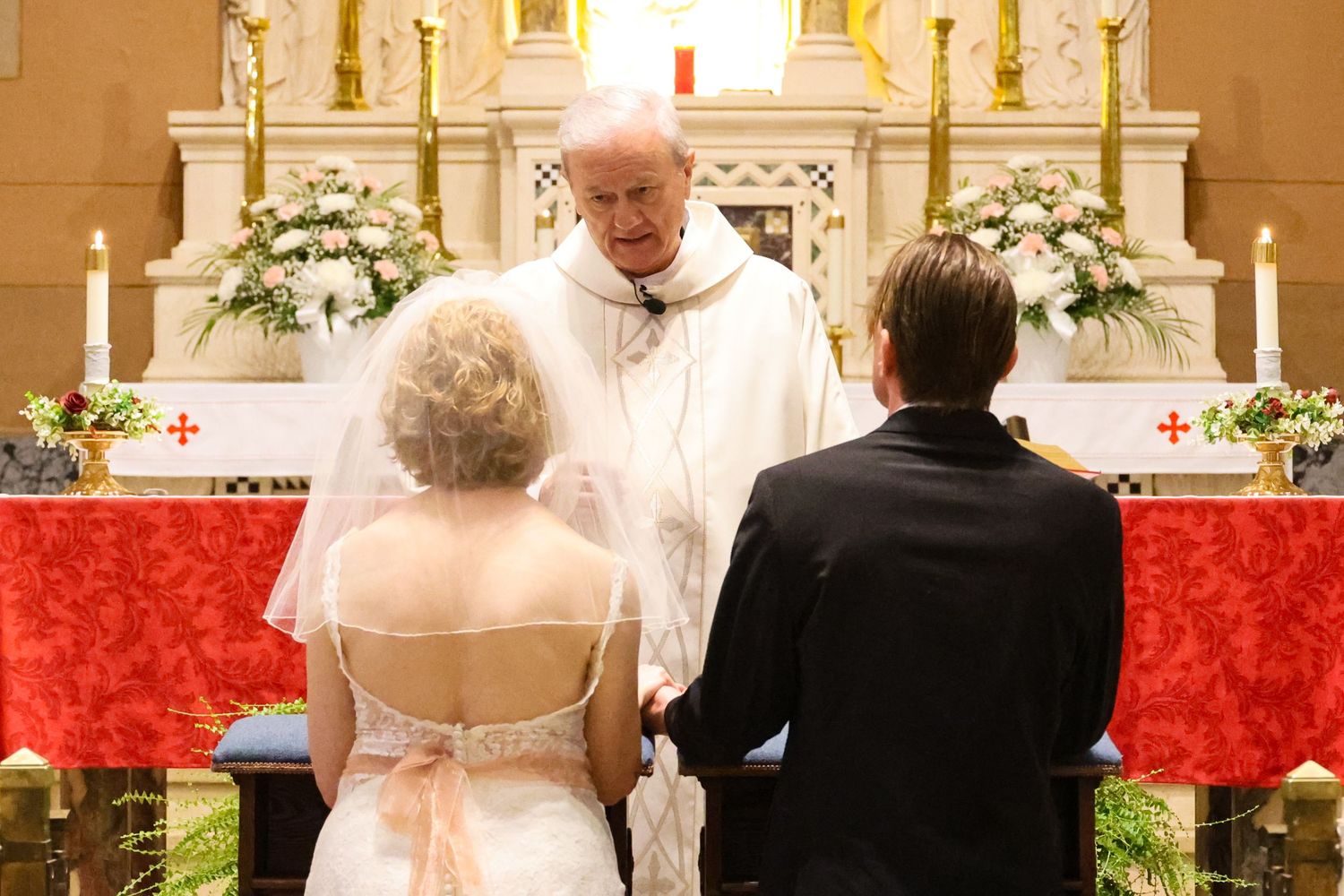 A church wedding ceremony with a bride and groom standing at the altar surrounded by white floral arrangements.