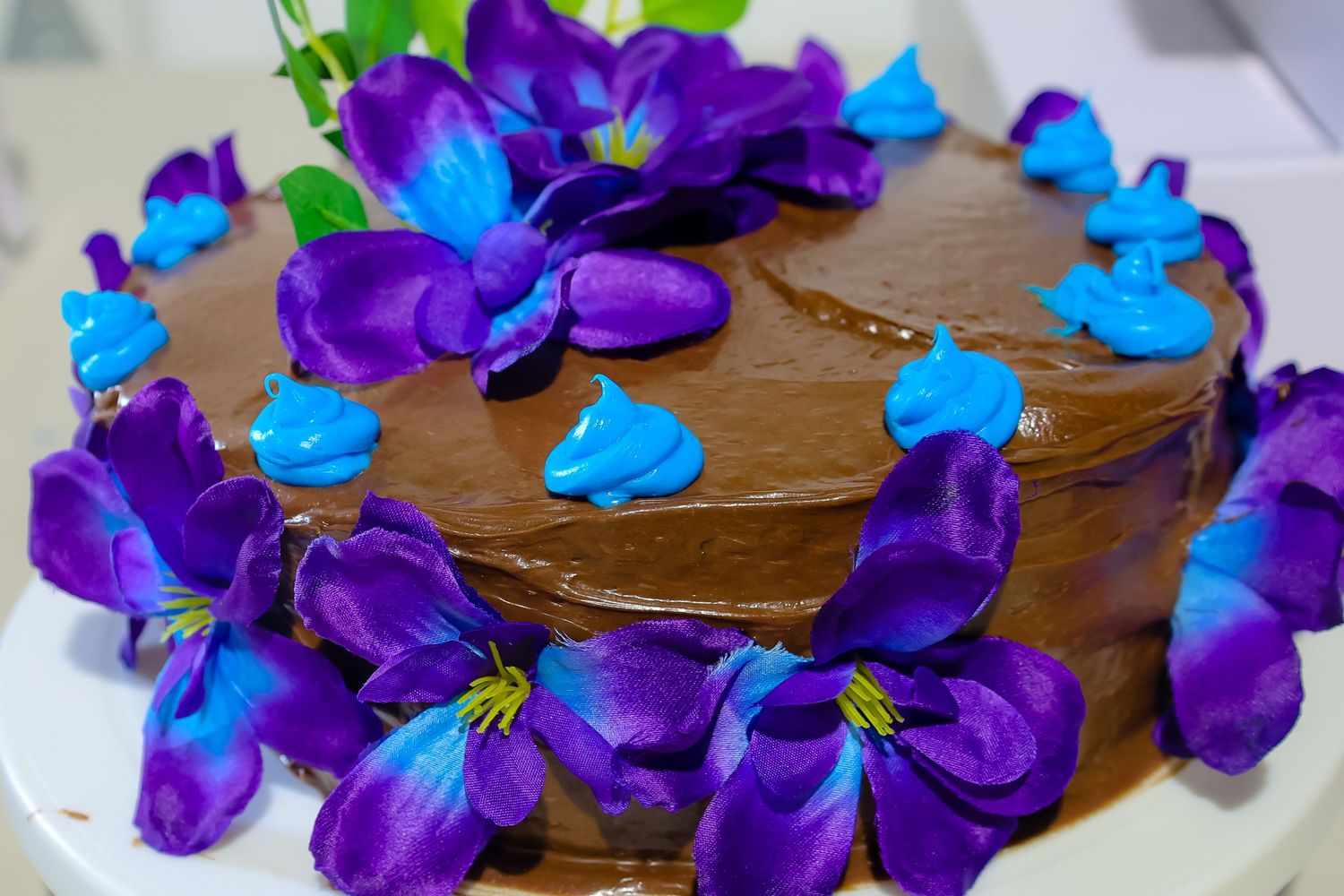 Chocolate frosted cake decorated with vibrant purple and turquoise flowers arranged in a beautiful pattern.