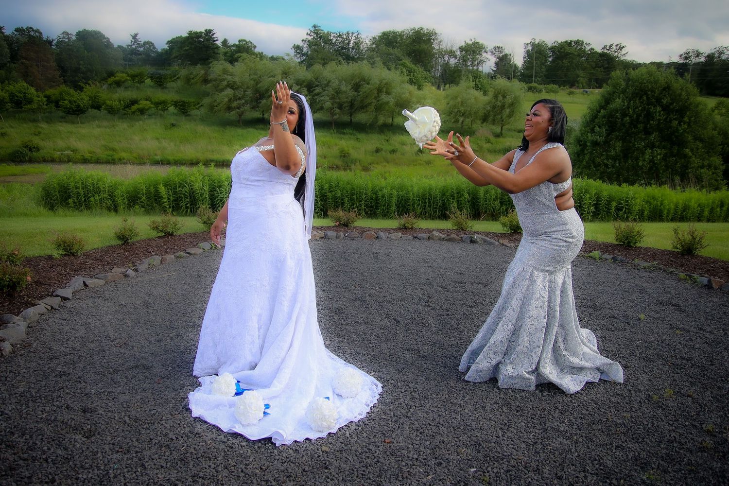 Two people in white wedding dresses pose playfully outdoors on a gravel path near green fields.