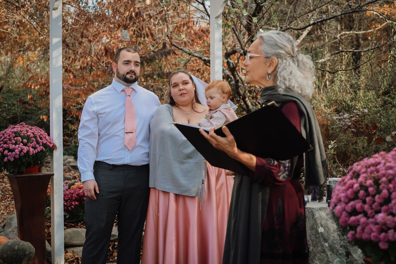 Wedding party poses together in autumn garden setting with pink mums and fall foliage in background.