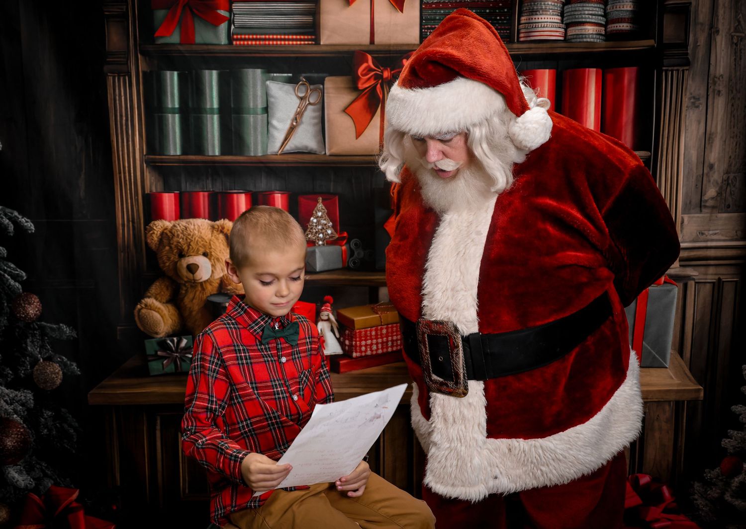 A child in plaid pajamas sits at a wooden table reading a letter while Santa Claus looks on during Christmas.