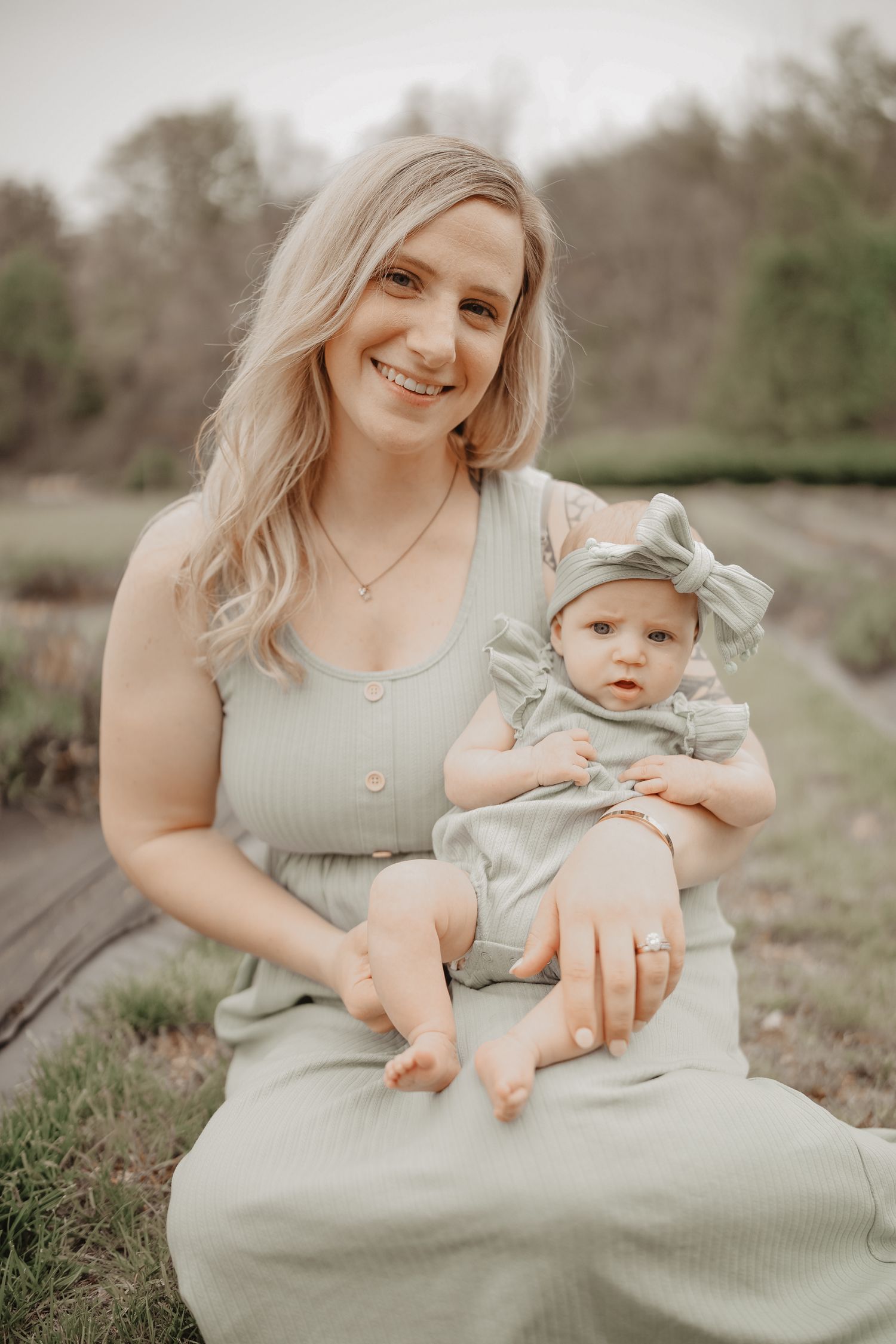 A tender outdoor portrait showing a parent and baby in matching soft sage green outfits sitting in a grassy field.