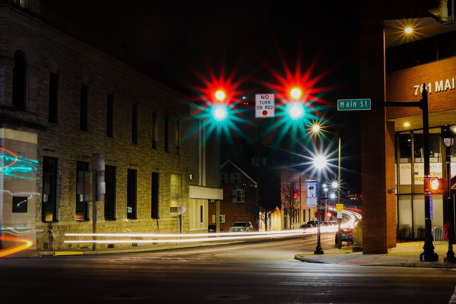 Night street scene in a small town with illuminated traffic lights glowing red against dark buildings and empty streets.
