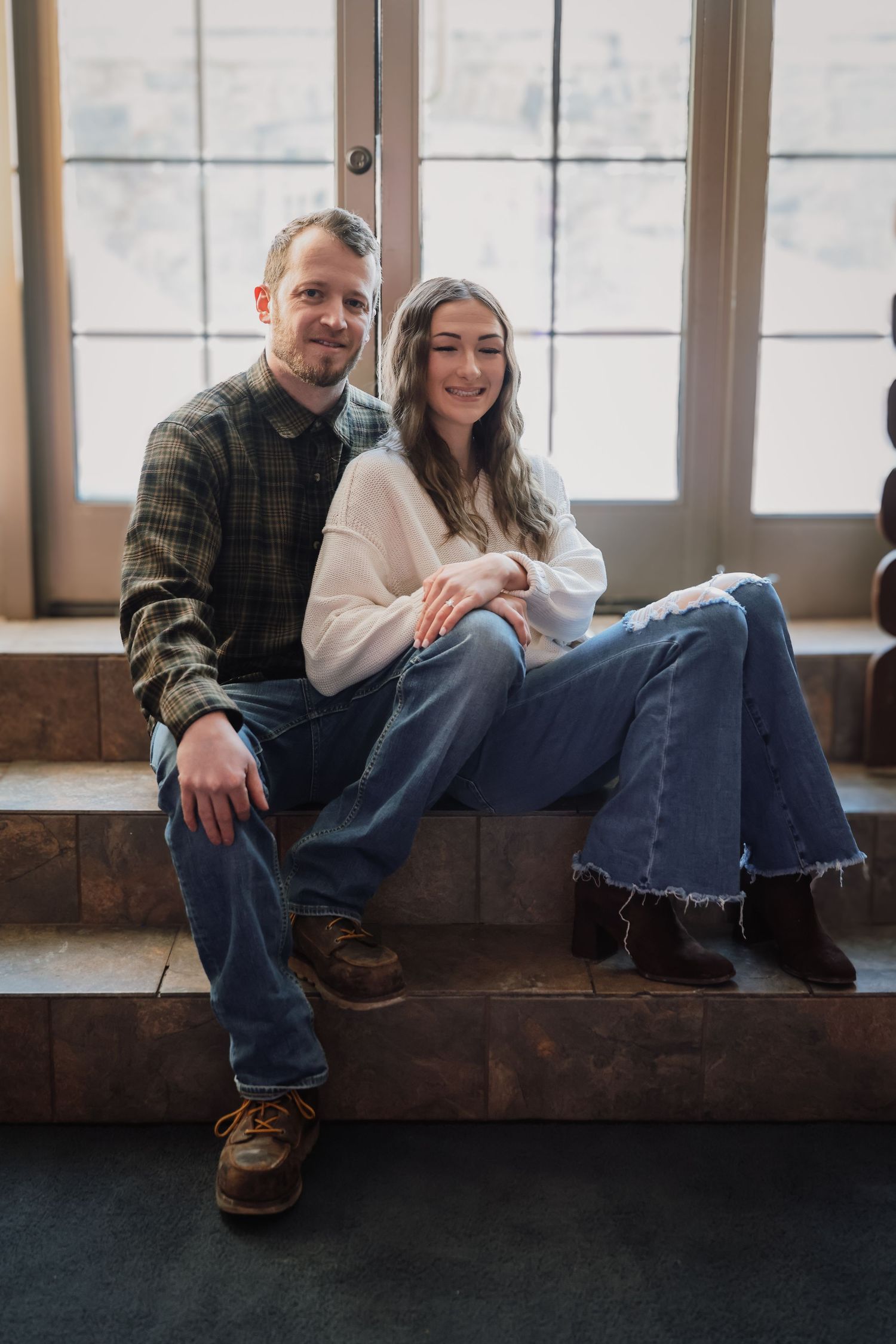 A couple sits together on a wooden bench near large windows in a casual indoor setting.