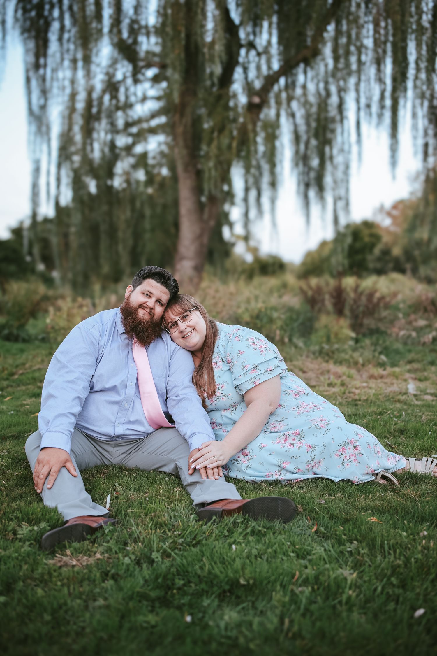 A couple relaxes together on green grass under hanging moss trees in a romantic outdoor setting.