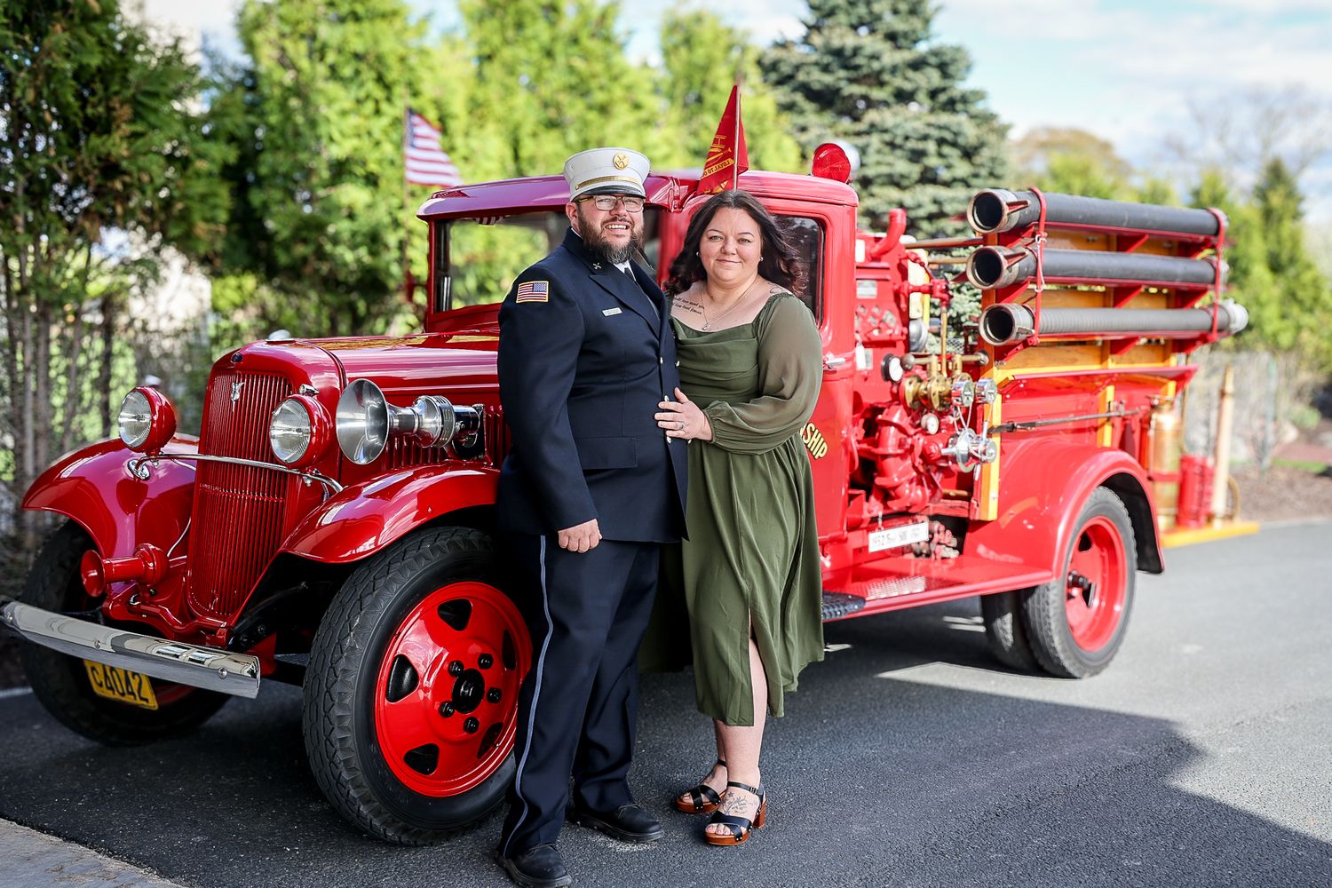 A couple poses in front of a vintage red fire truck, man in uniform and woman in green dress, outdoors.