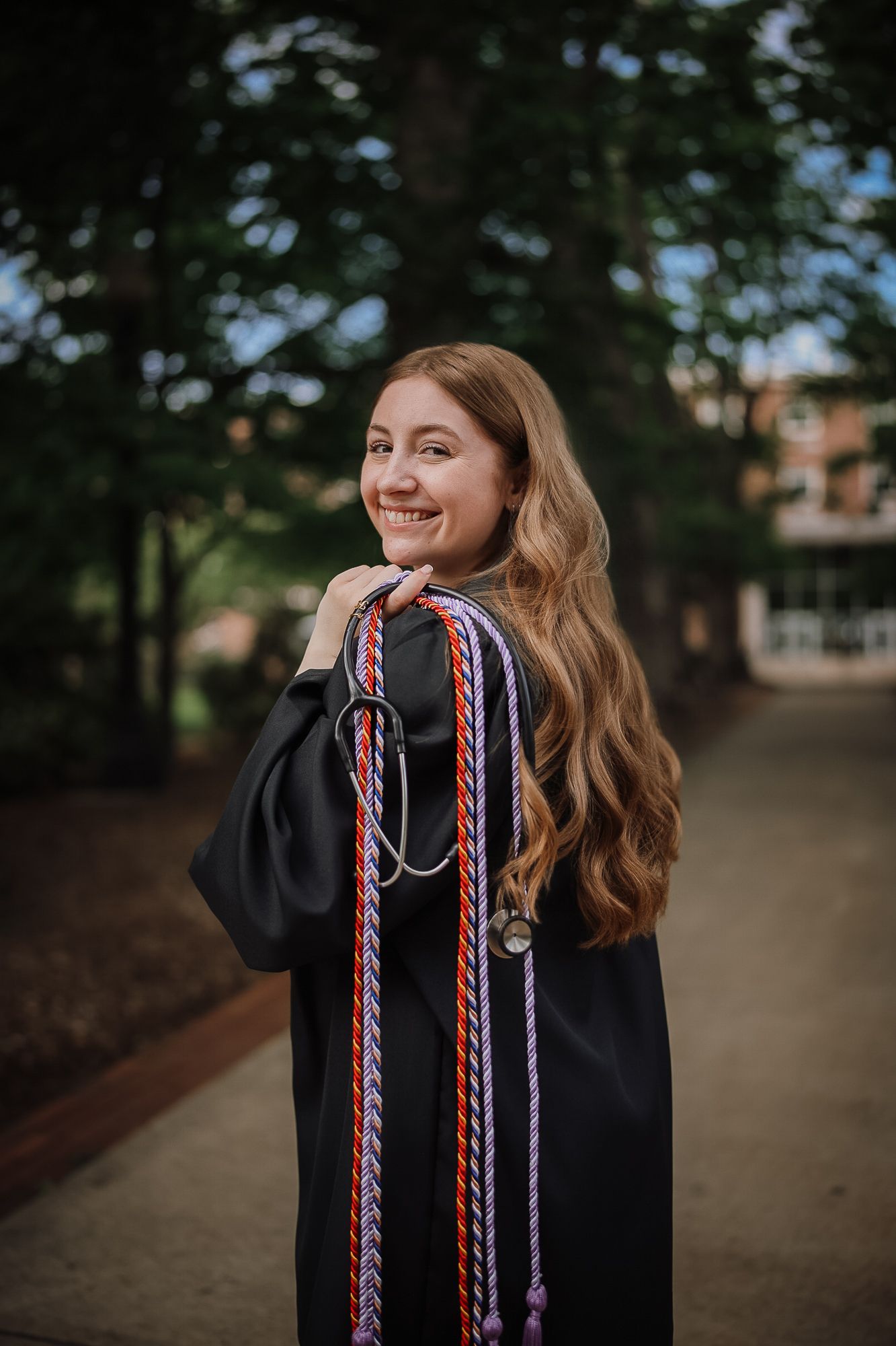Smiling young woman in black graduation gown holding colorful honor cords outdoors on a tree-lined campus path.
