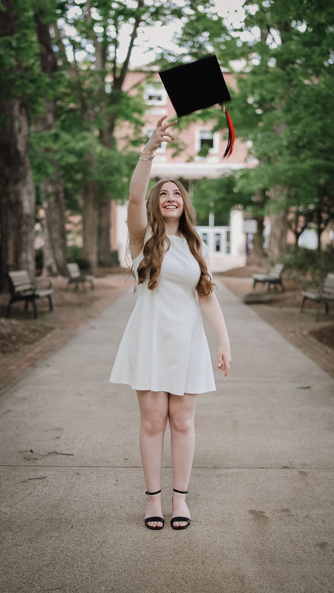 Smiling graduate in white dress tossing black cap in tree-lined campus walkway, celebrating graduation day.