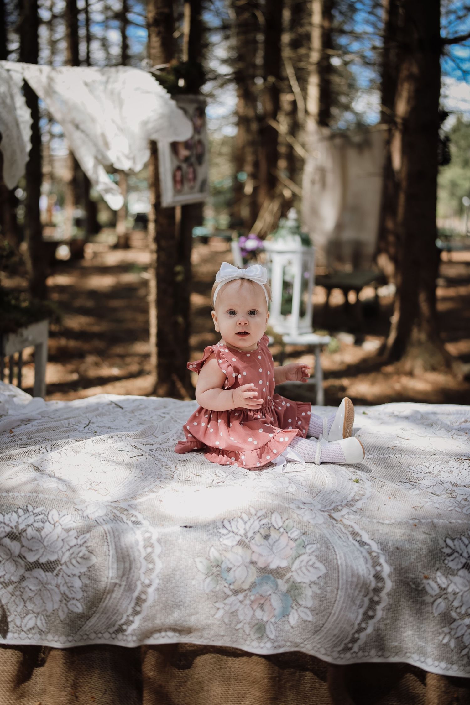 Baby girl in pink polka dot dress and bow headband sitting on a floral quilt outdoors in a sunlit forest setting.