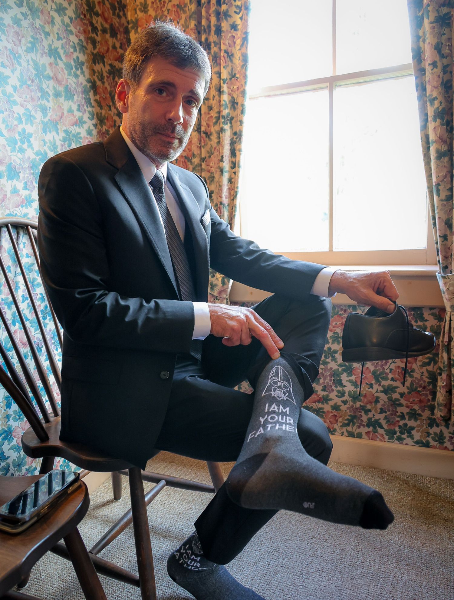 Man in suit sitting on chair displaying Star Wars 'I Am Your Father' novelty socks while holding a shoe.