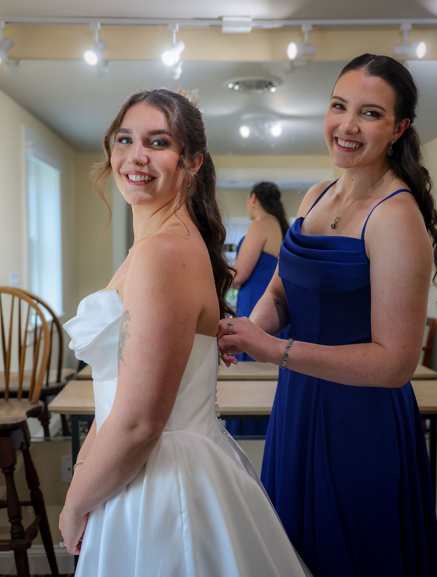 Bridesmaid in royal blue dress buttoning the back of a bride's white wedding gown, both smiling in a bright room.