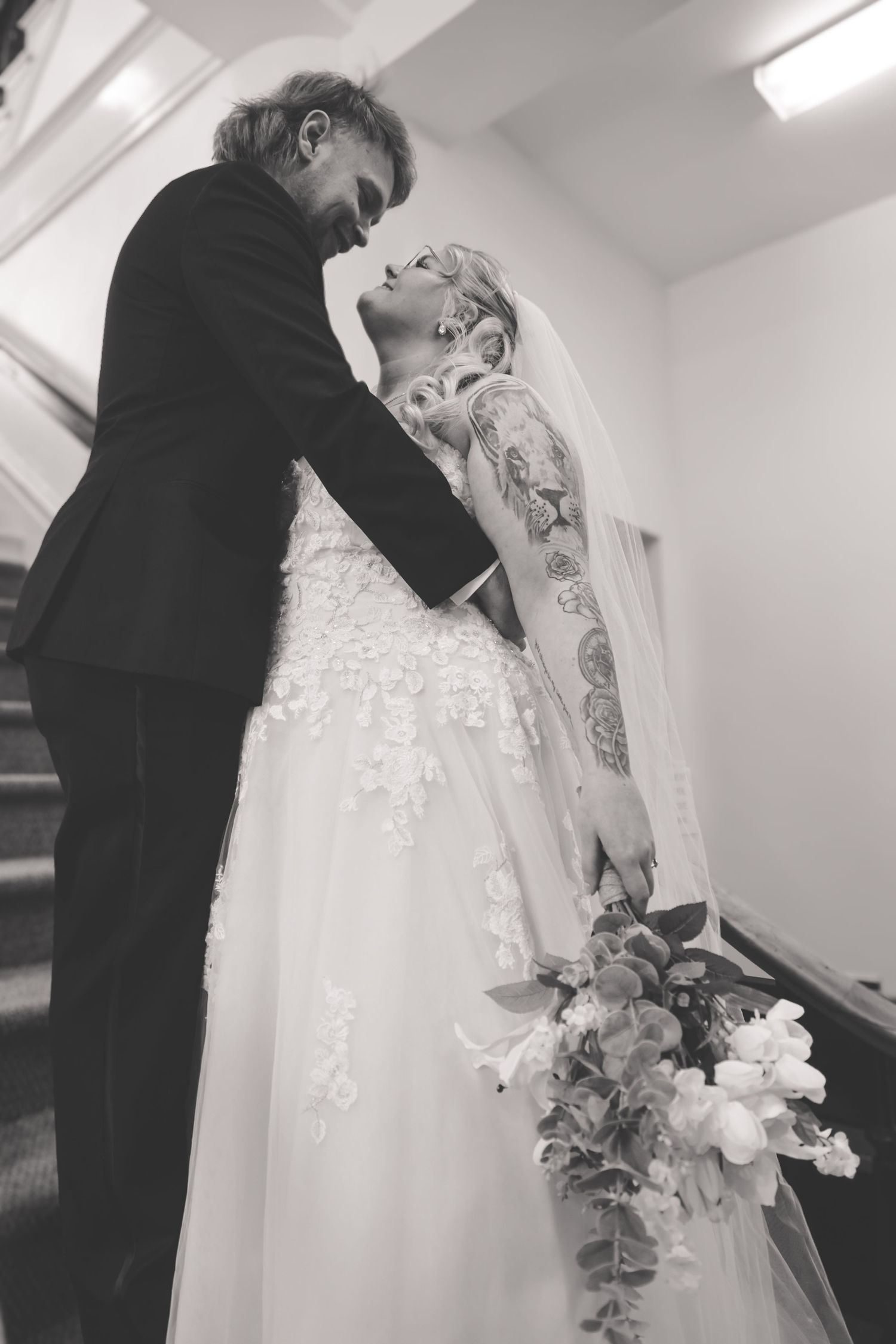 Black and white photo of tattooed bride in lace gown holding bouquet, leaning toward groom in suit on staircase.