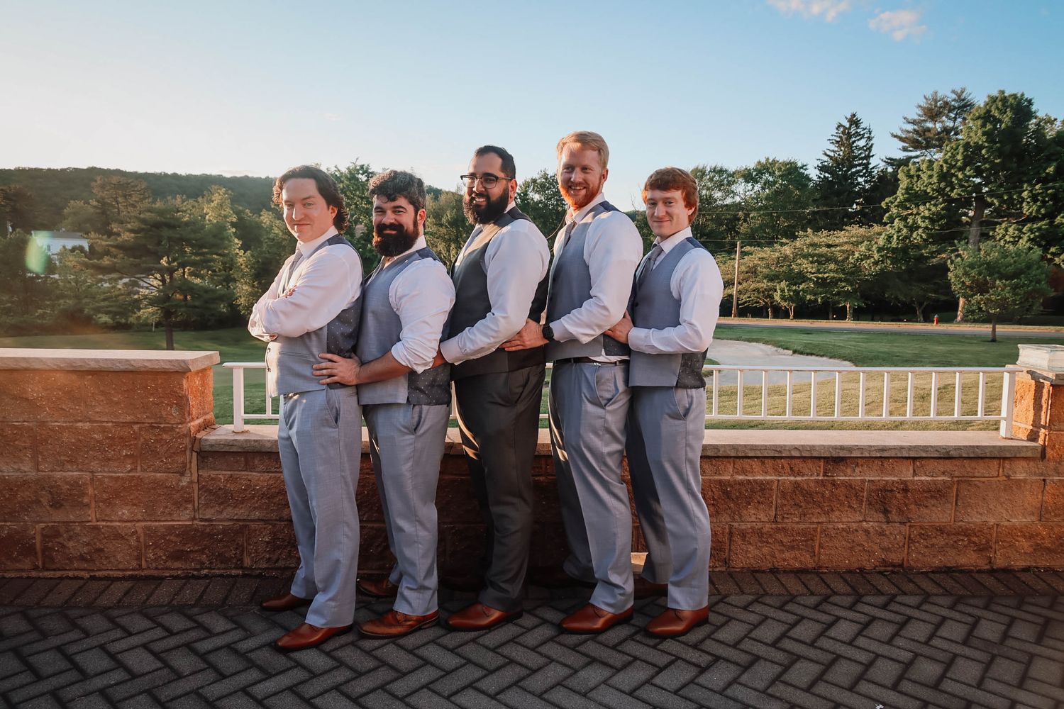 Five groomsmen in gray vests and trousers pose confidently on an outdoor terrace at a wedding.