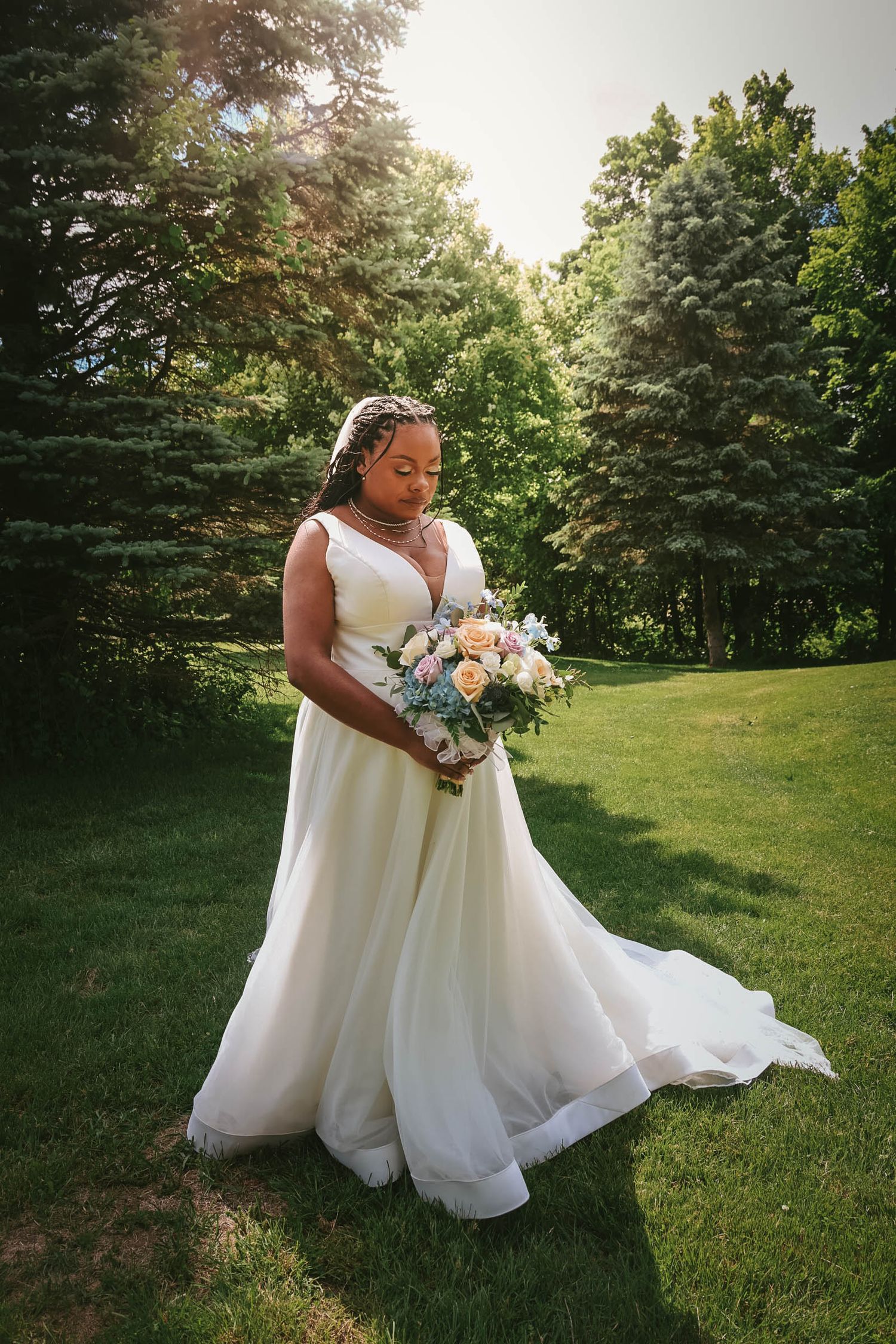 A bride in a flowing white gown holds a pastel bouquet while standing on lush green grass surrounded by tall trees.