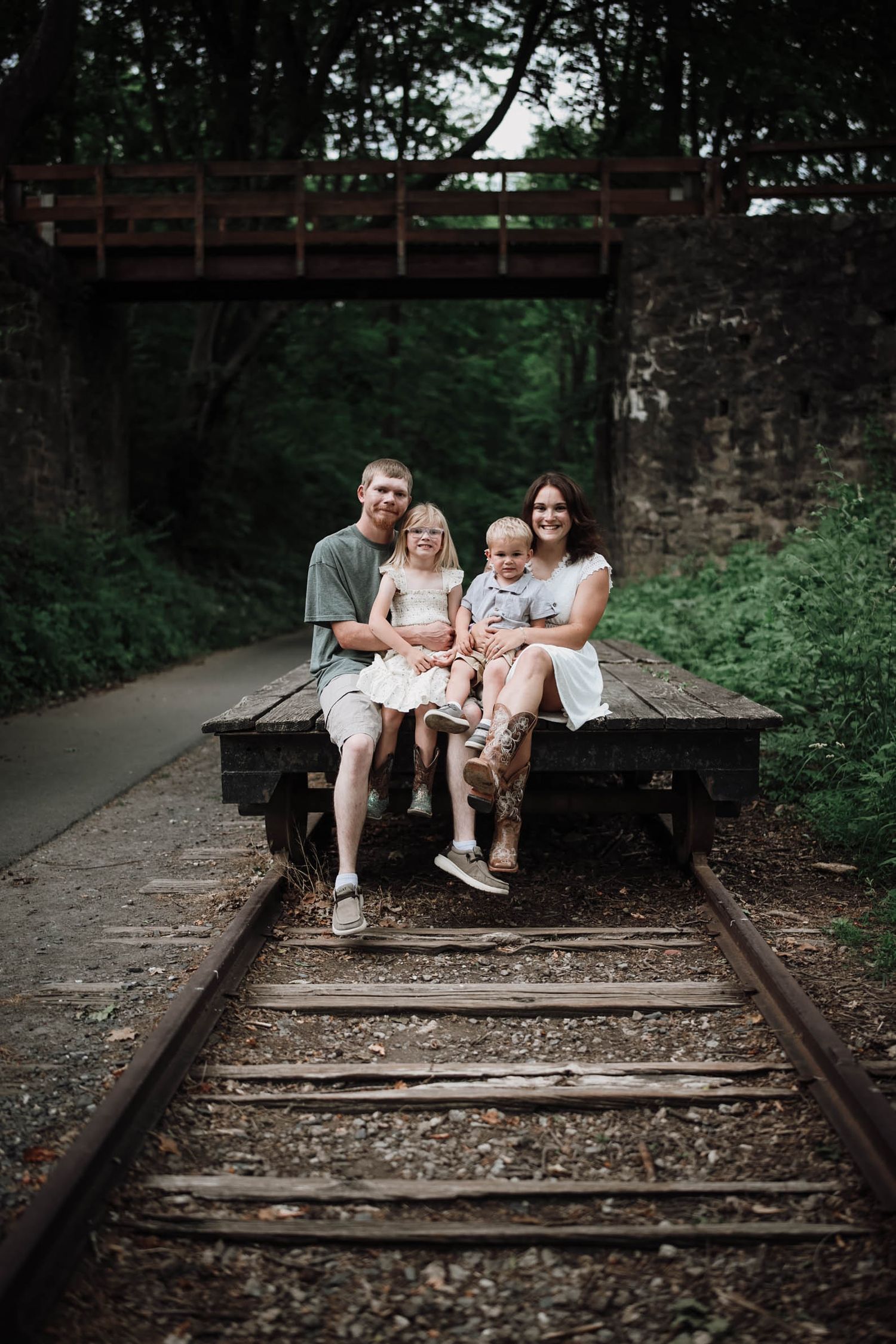 Family of four sitting on a vintage railroad flatcar on train tracks surrounded by lush greenery.