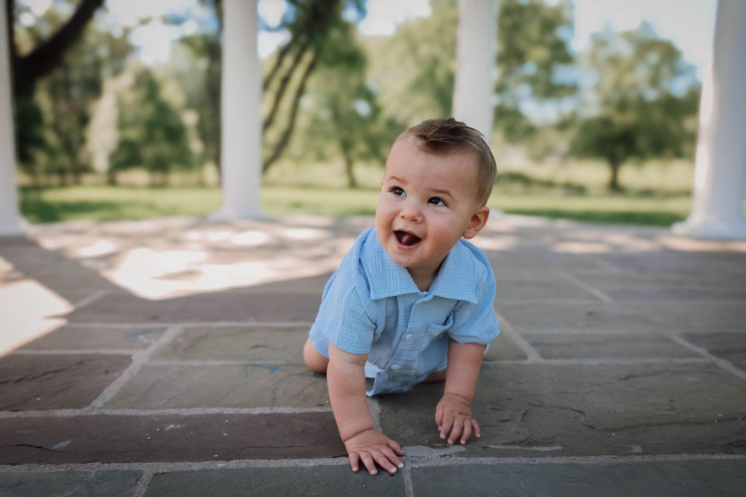 A happy baby boy in a blue romper crawls on a brick patio surrounded by white columns and green trees.