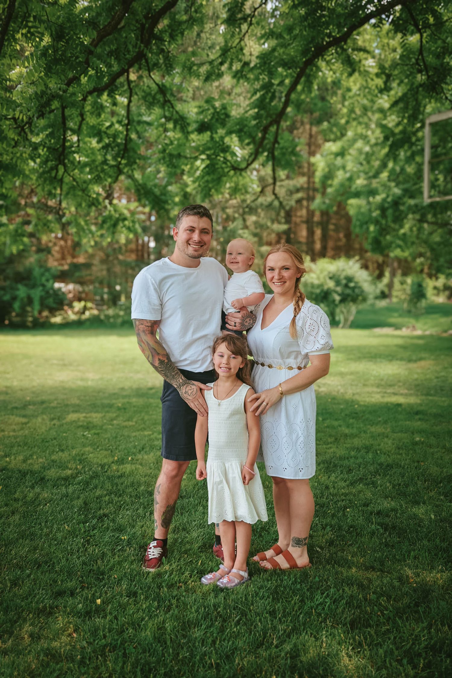 Family of four posing outdoors on green grass under trees, parents with tattooed arms holding baby and young girl in white dress.