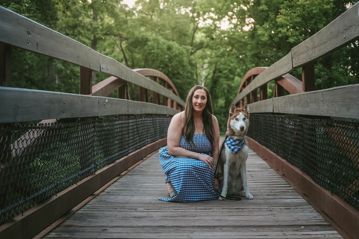 Woman in blue maxi dress kneeling beside Husky dog on wooden bridge in forest park.