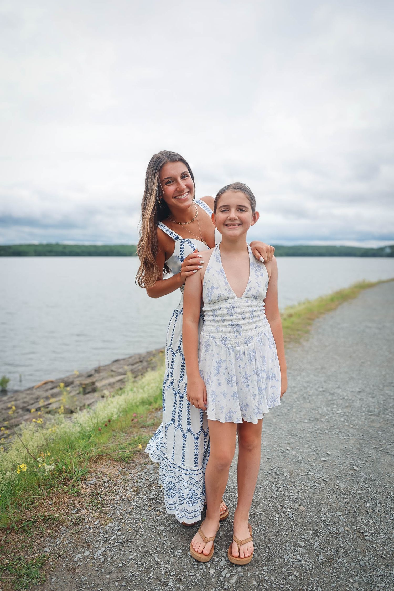 Two girls in summer dresses pose together on a lakeside path under a cloudy sky.