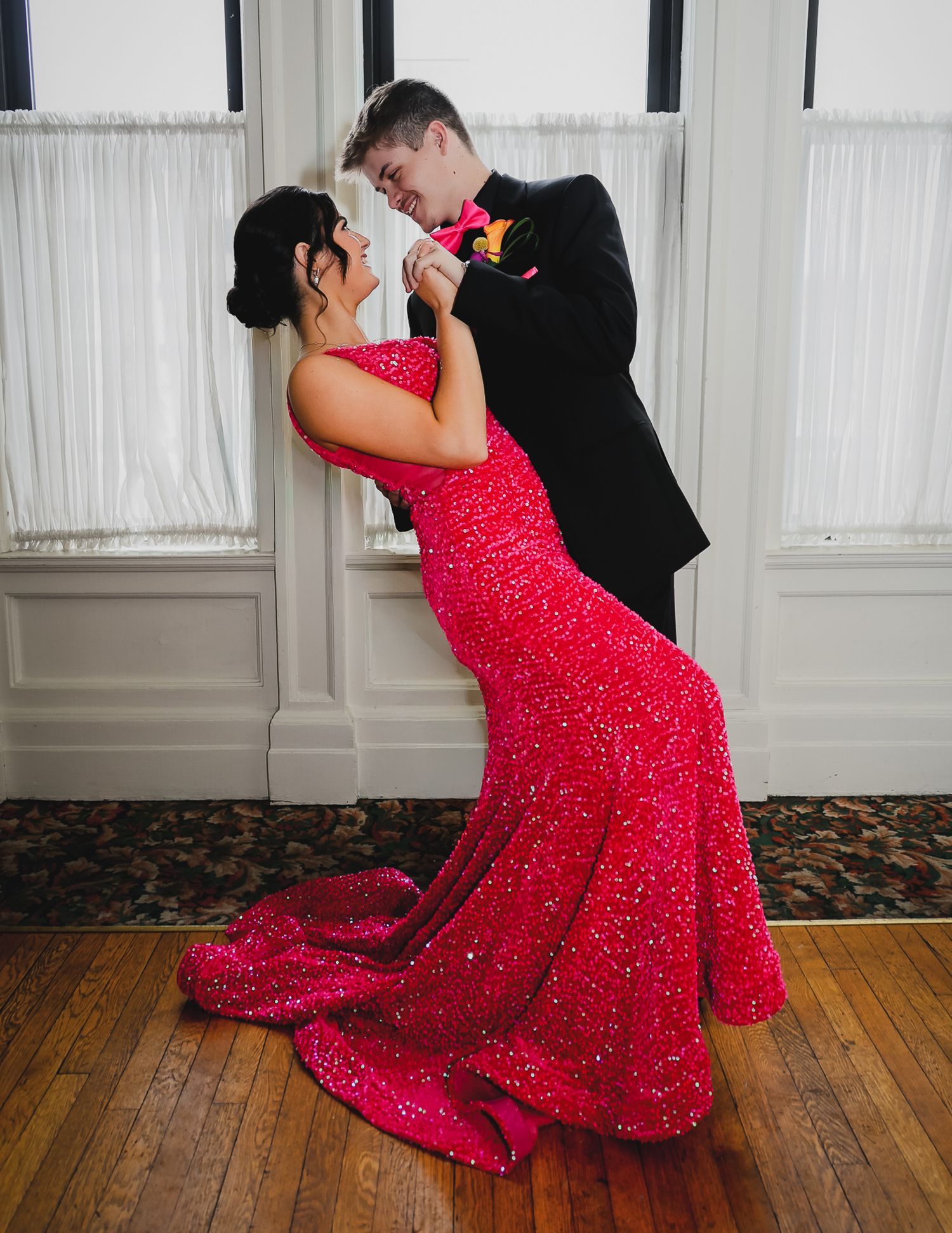 Couple dancing at prom, woman in red sequin mermaid gown, man in black tuxedo with floral boutonniere.