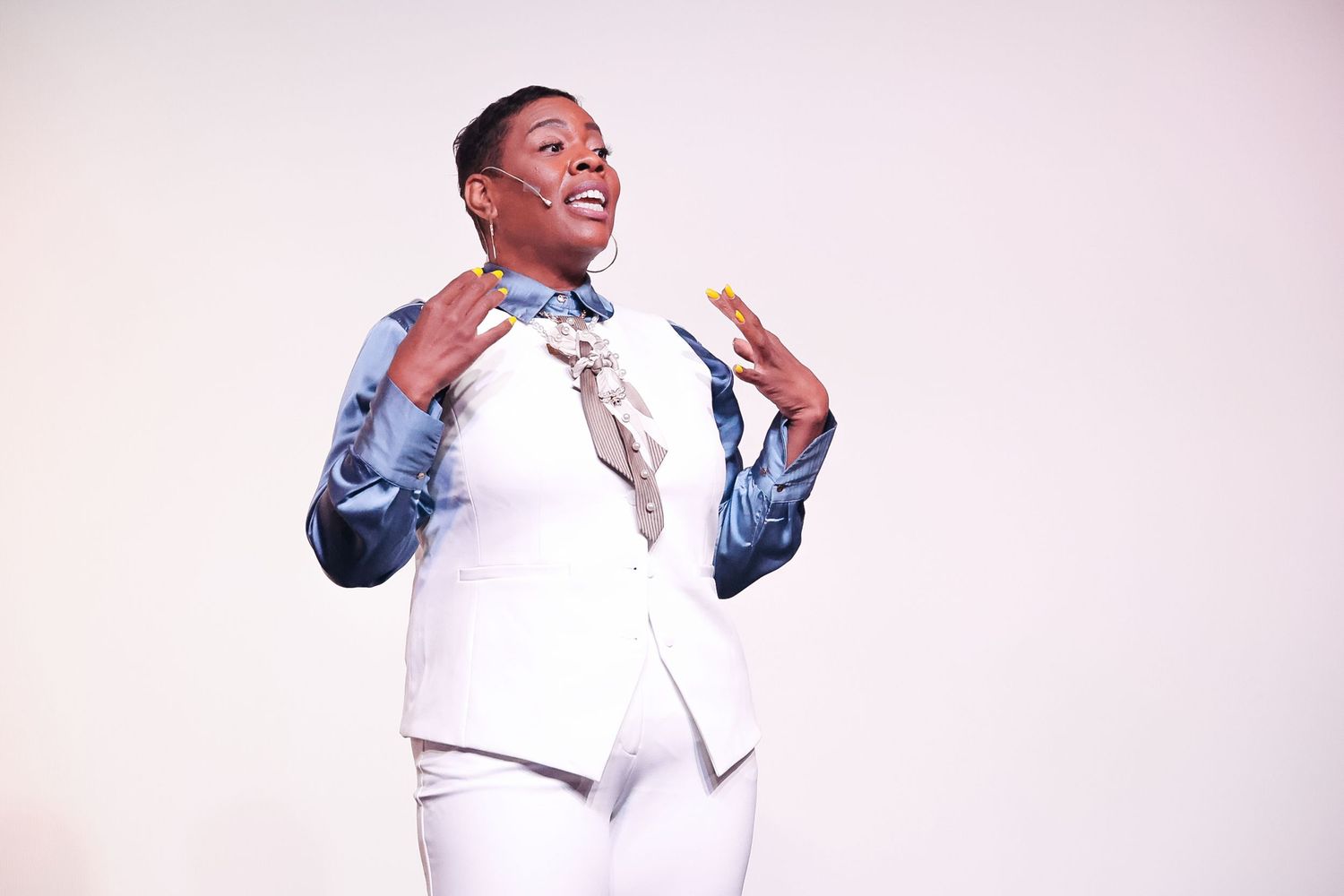 A confident Black woman in a white suit gestures expressively while speaking on a bright stage.