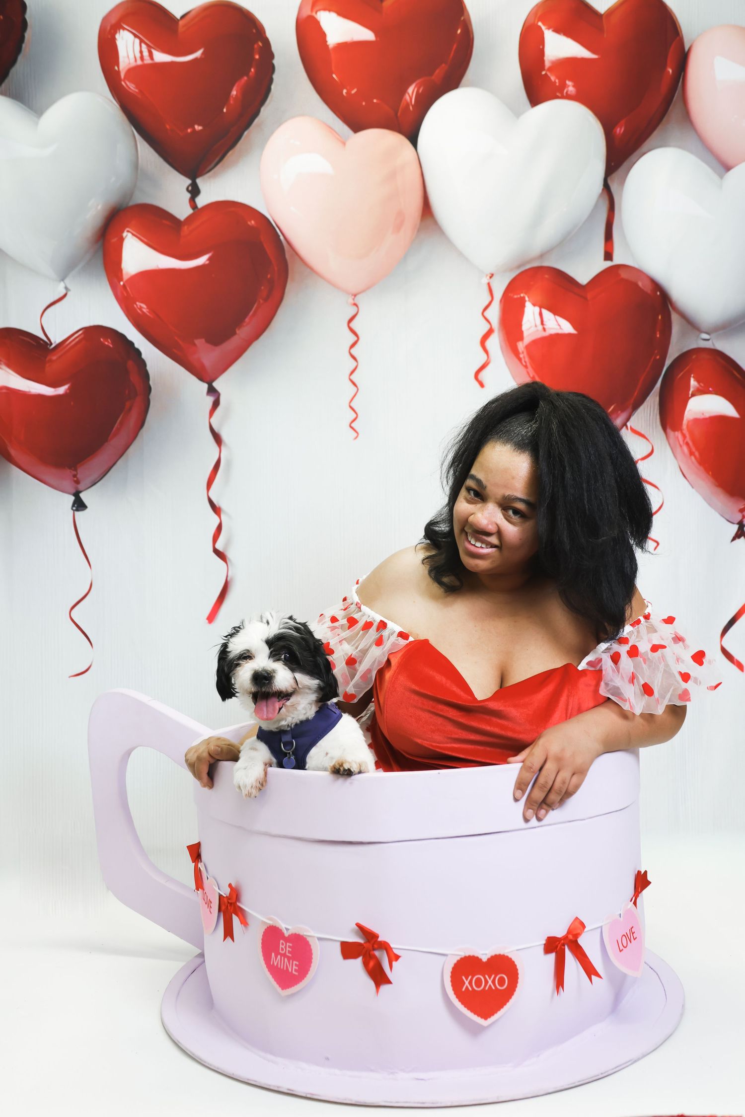 A smiling woman in a red dress poses in a giant teacup with her dog, surrounded by heart-shaped balloons for Valentine's Day.
