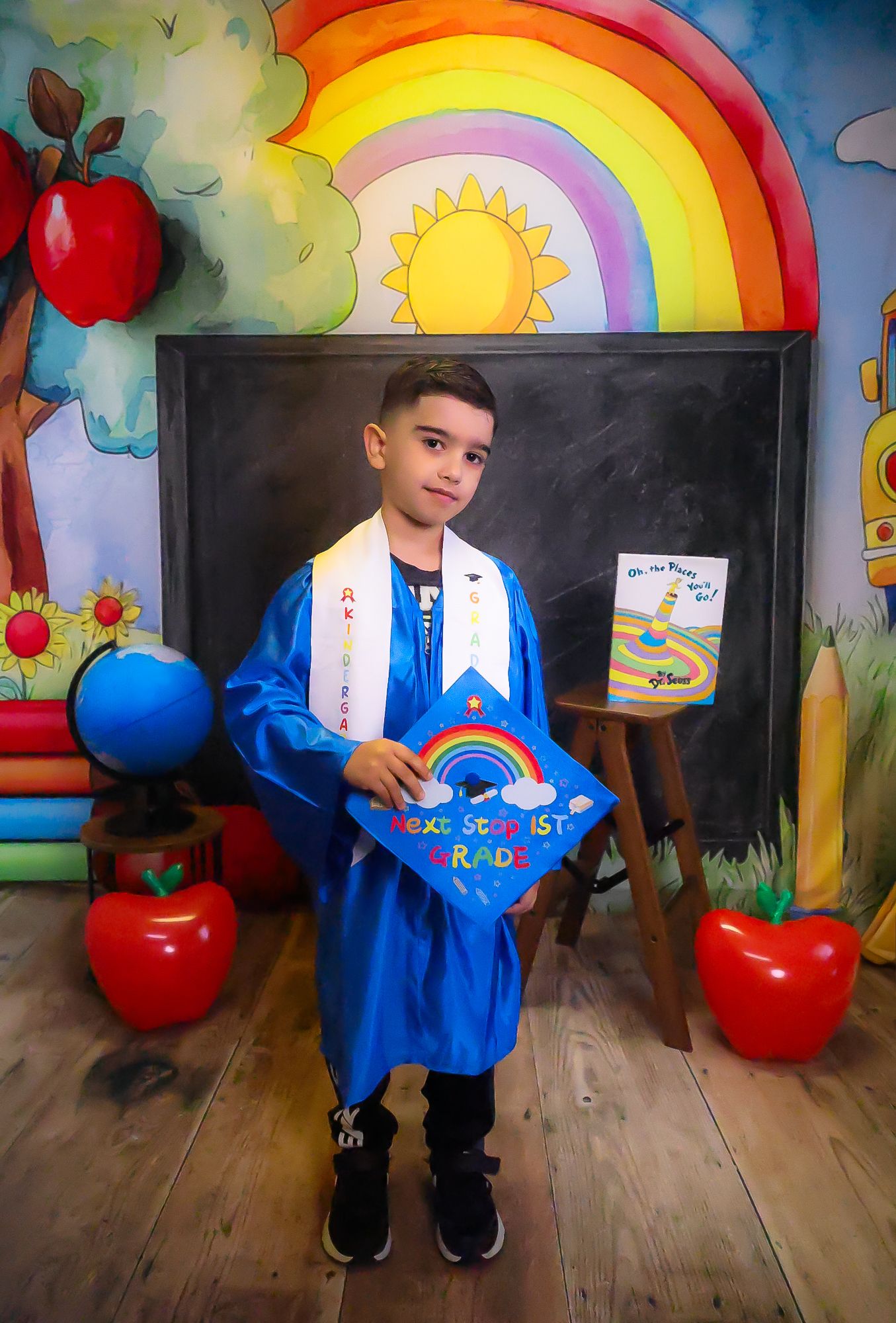 Young boy in blue graduation gown and cap holding diploma, posing in front of colorful rainbow backdrop.