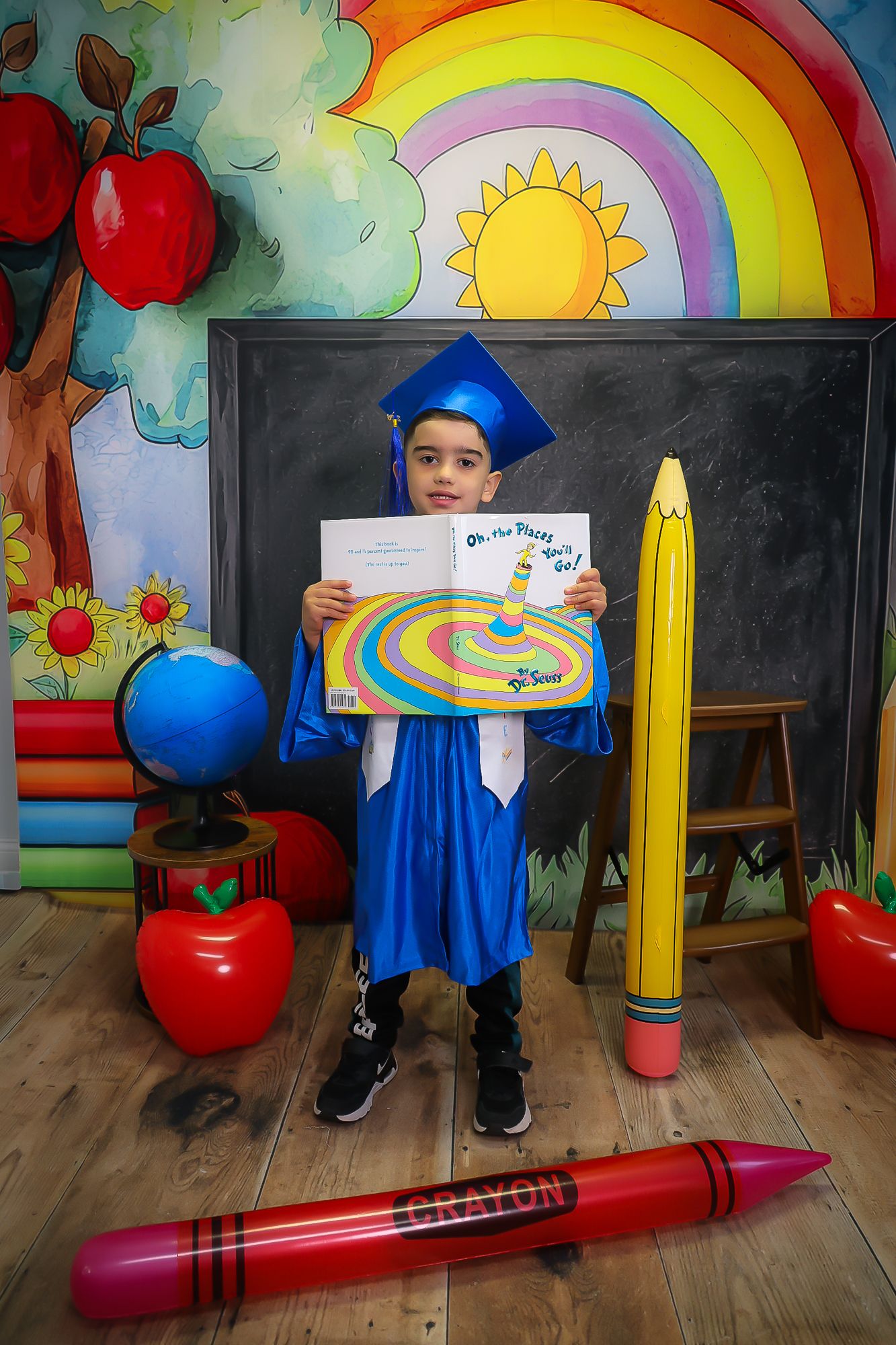Young boy in blue graduation cap and gown holds Dr. Seuss book in colorful school-themed photo backdrop.