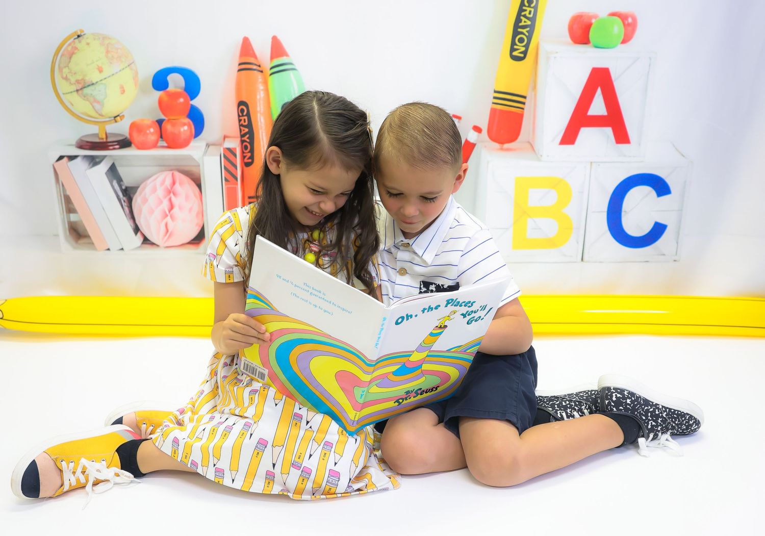 Two young children sit together reading a colorful book, surrounded by ABC letters and school supplies.